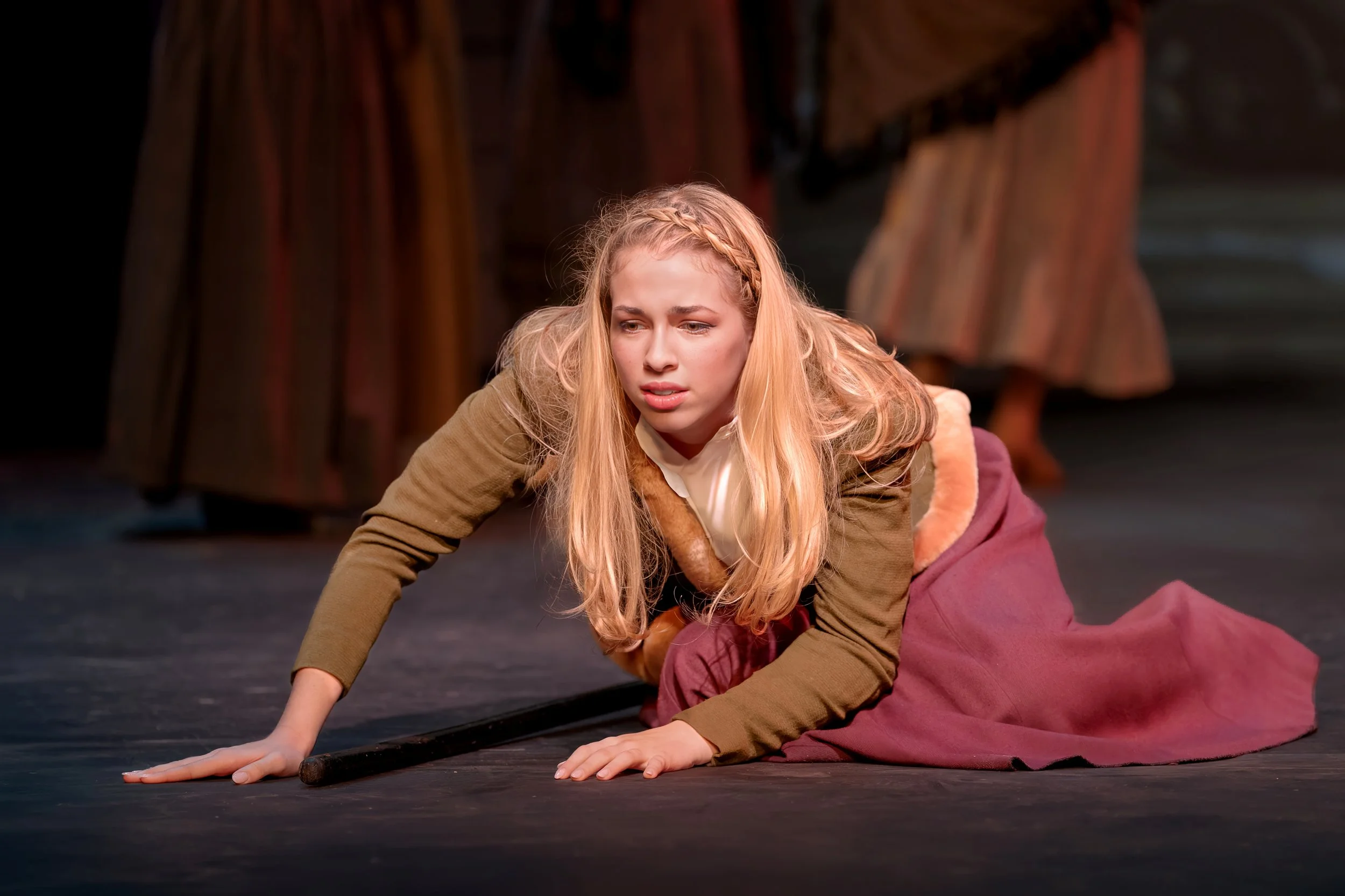 Young woman with long blonde hair, wearing a brown jacket and a cream-colored shirt, on her hands and knees on a stage, with a dark background and another person partially visible behind her.