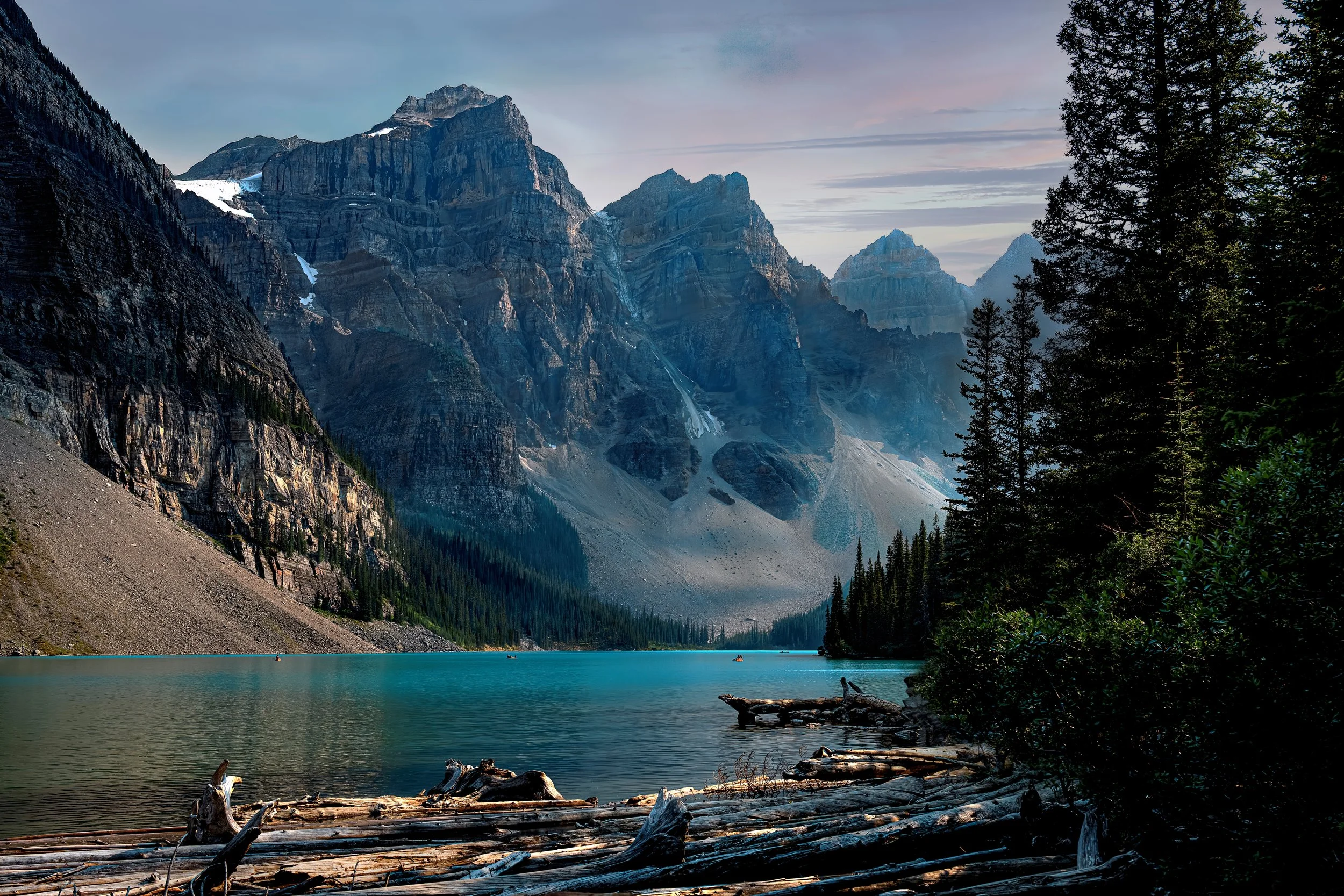 Scenic view of a mountain lake with turquoise water, surrounded by tall evergreen trees and rugged snow-capped peaks in the background, with a partly cloudy sky.