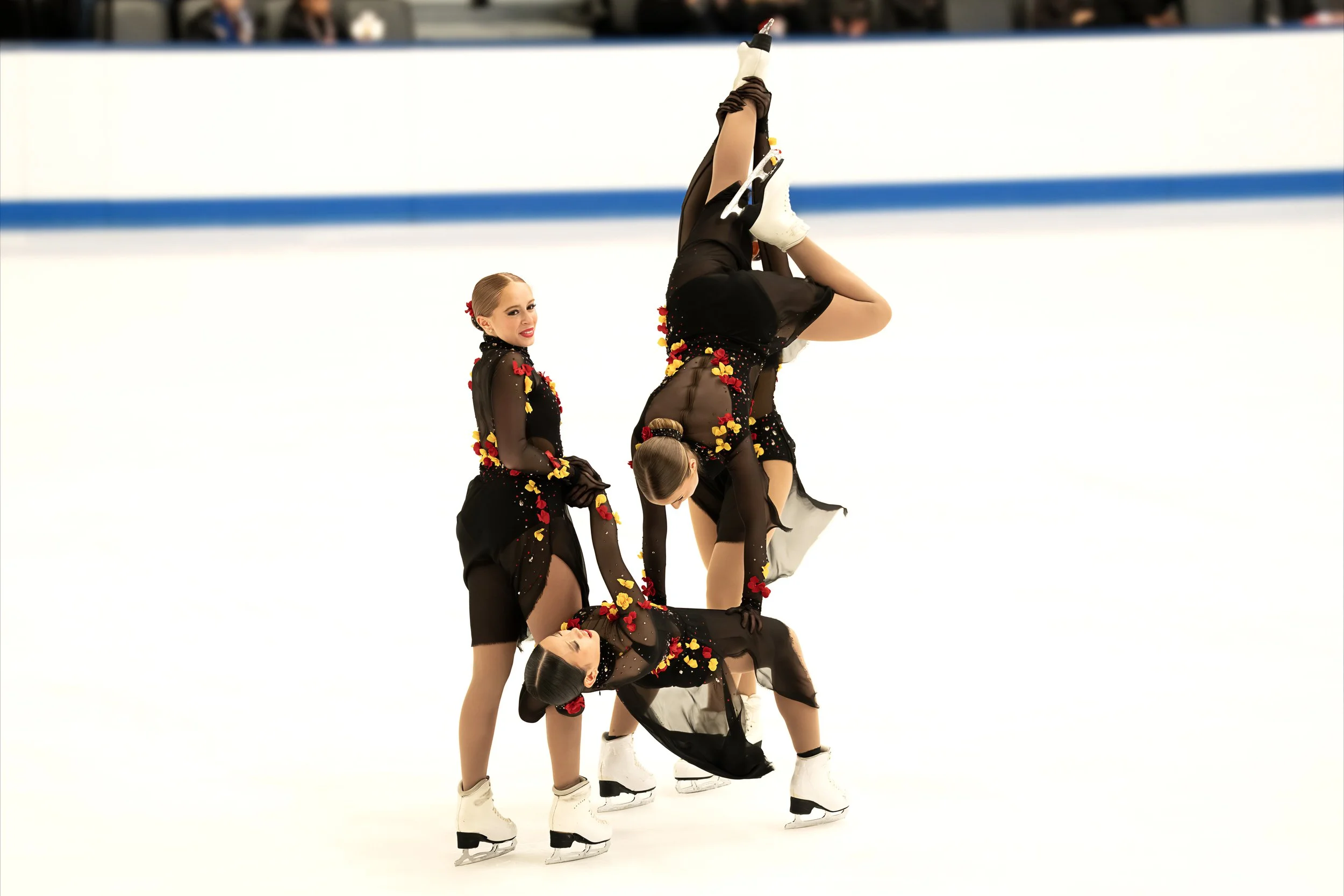 Six figure skaters dressed in black costumes with yellow and red flower designs perform an acrobatic skating routine on the ice rink, with some skaters balancing upside down while others support them.