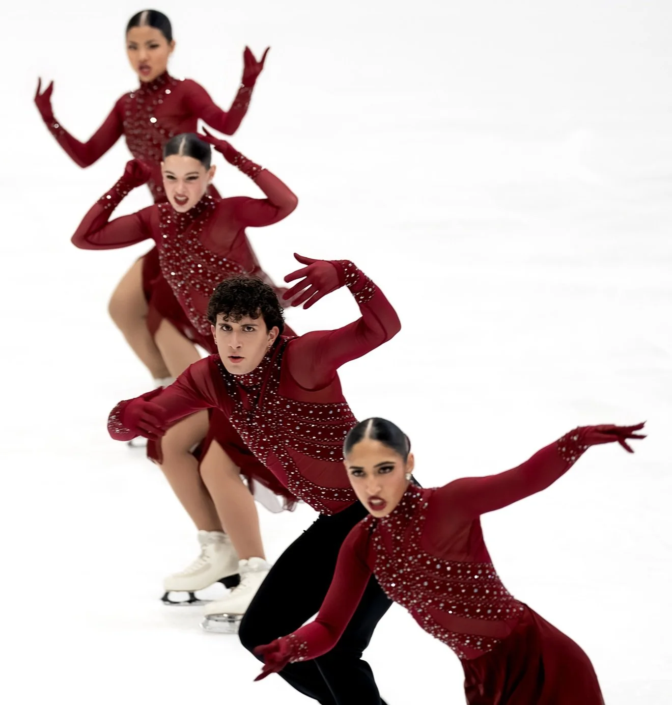 Four figure skaters wearing matching red costumes perform a synchronized routine on ice, with arms raised and poses aligned.