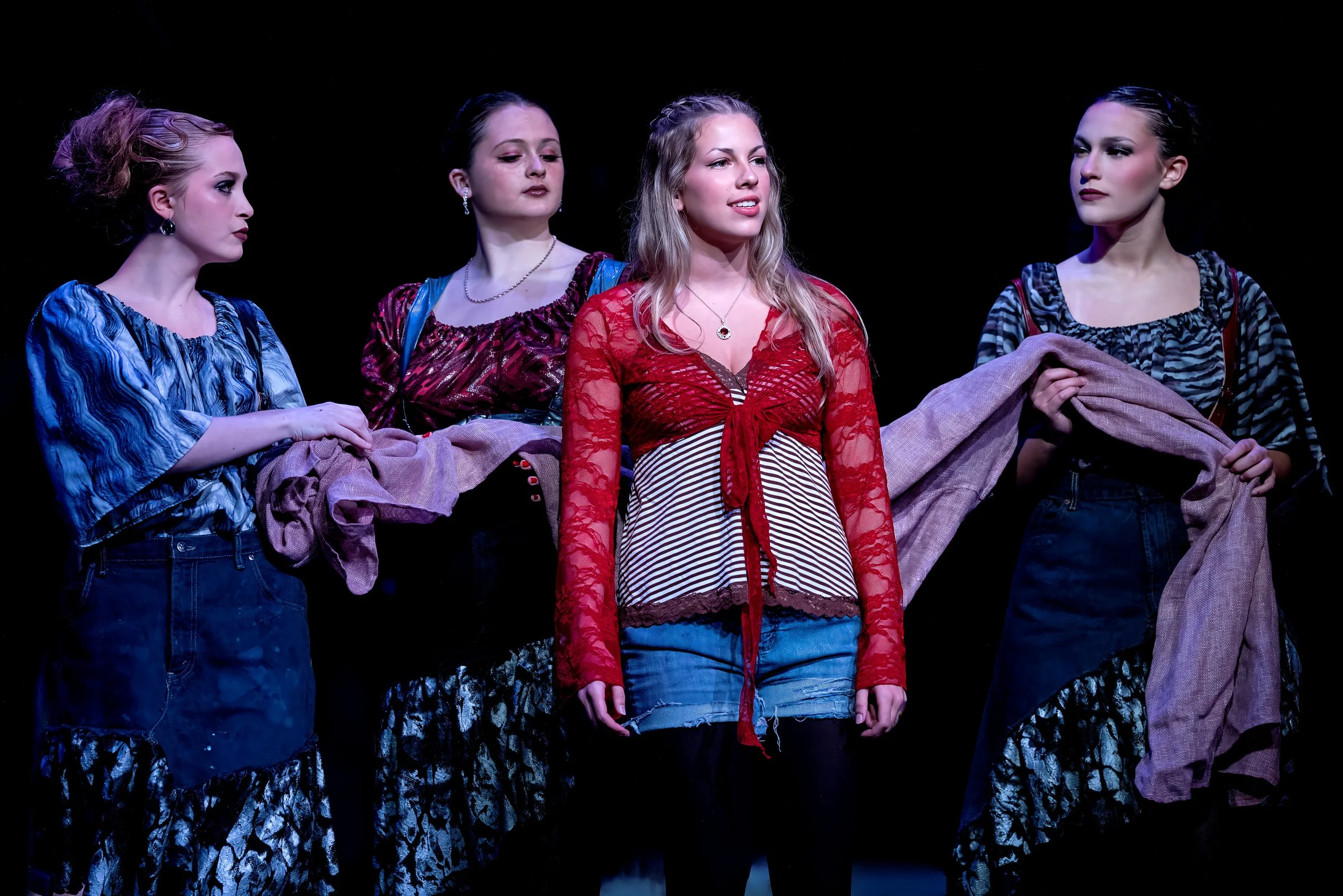 Four young women stand on stage, dressed in colorful clothing, holding a large piece of fabric, with black background.