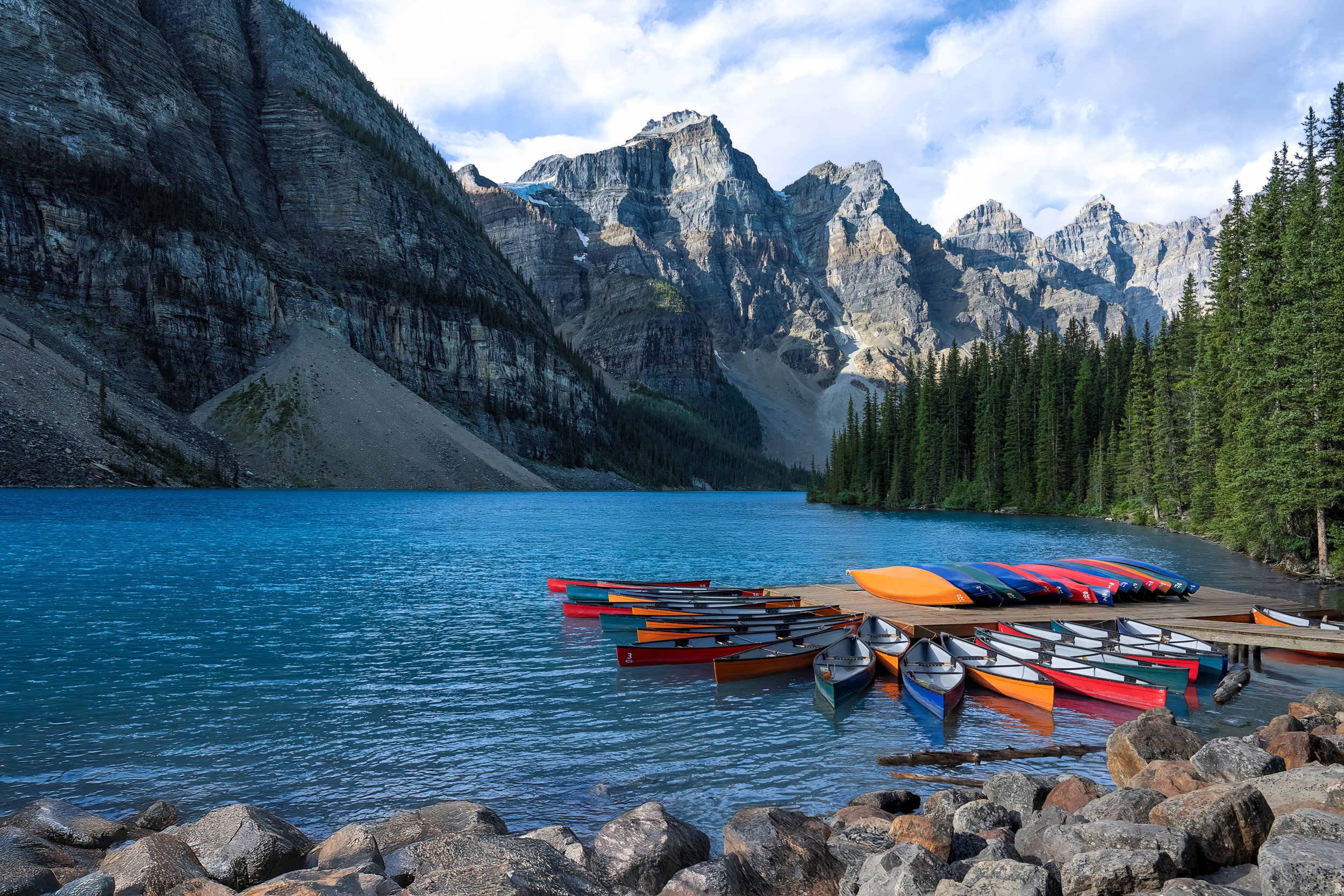 A scenic mountain lake with a dock of colorful canoes and kayaks, surrounded by forested shoreline and towering rocky mountains under a partly cloudy sky.