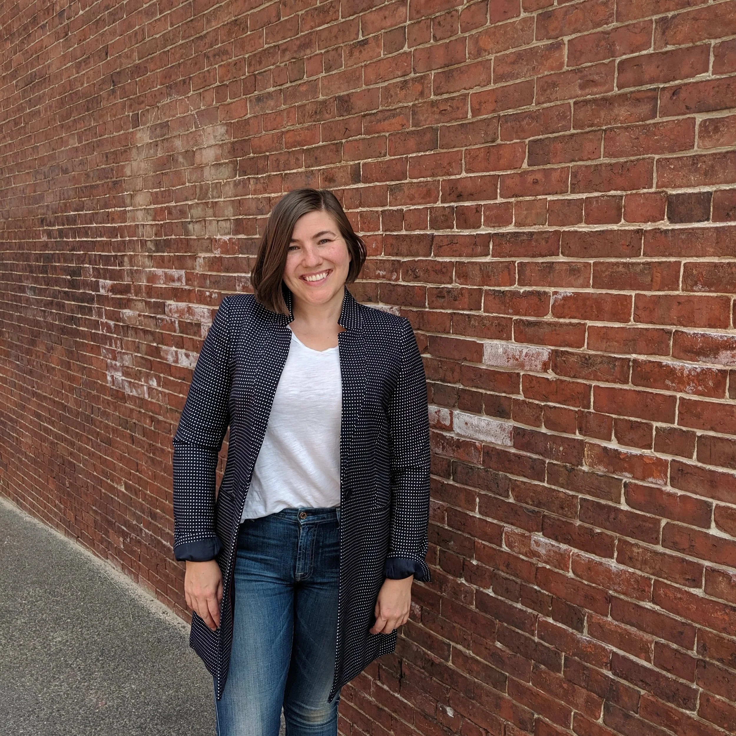 A woman with shoulder-length brown hair smiling, wearing a polka dot blazer and blue jeans, standing against a red brick wall.