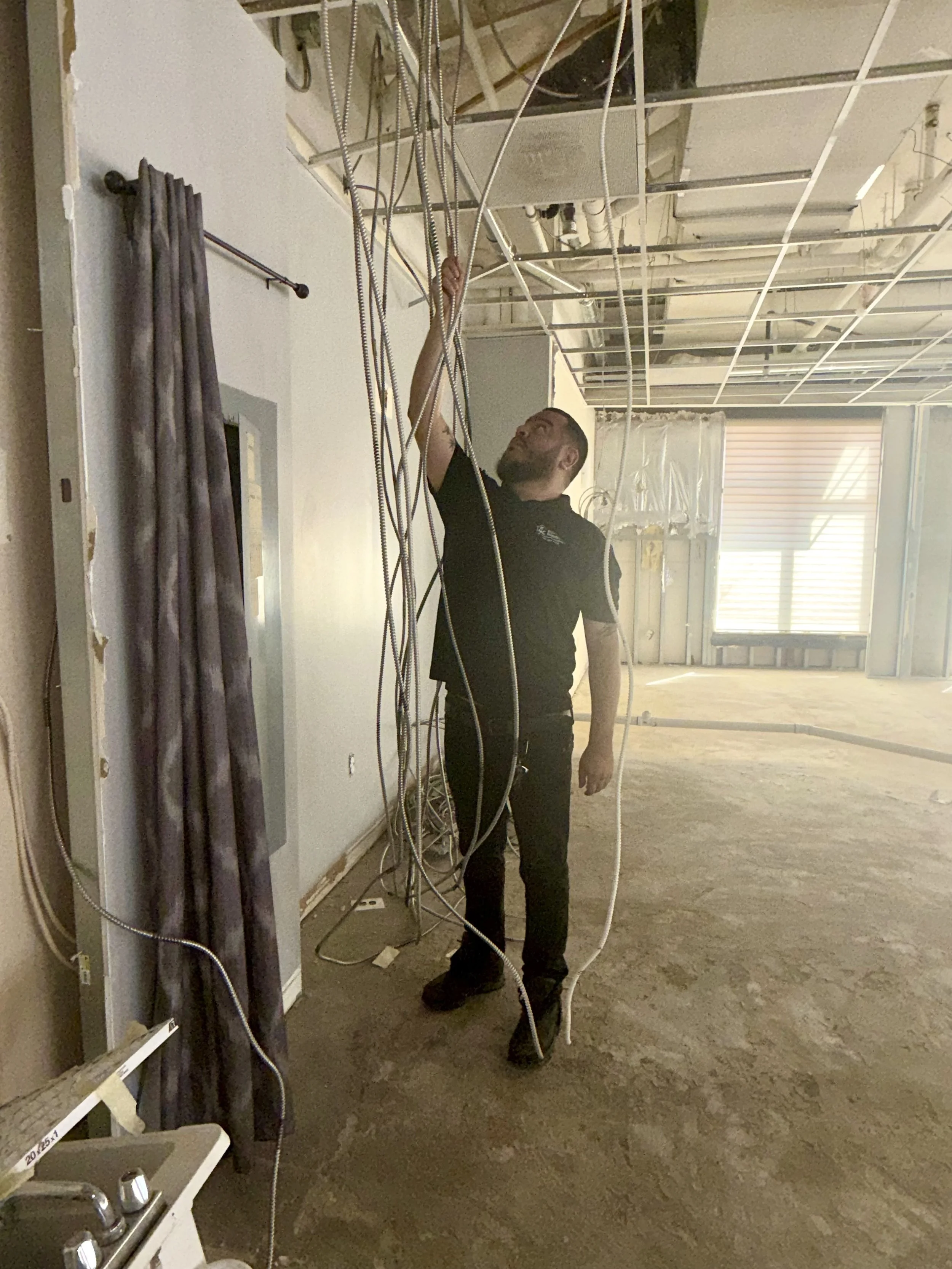A man working on exposed ceiling infrastructure, wires, and piping in a demolished shell for a tenant fit out.