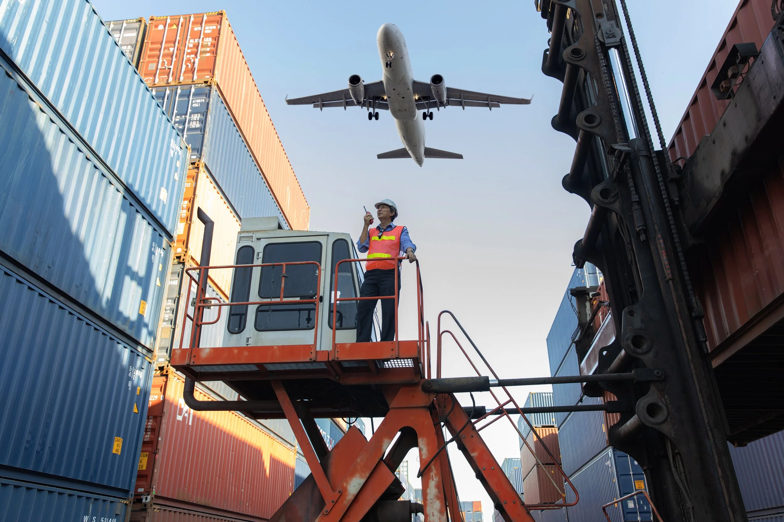 Un trabajador en una grúa aérea con un chaleco de seguridad naranja y un casco azul, inspecciona un avión de carga que pasa en un aeropuerto.
