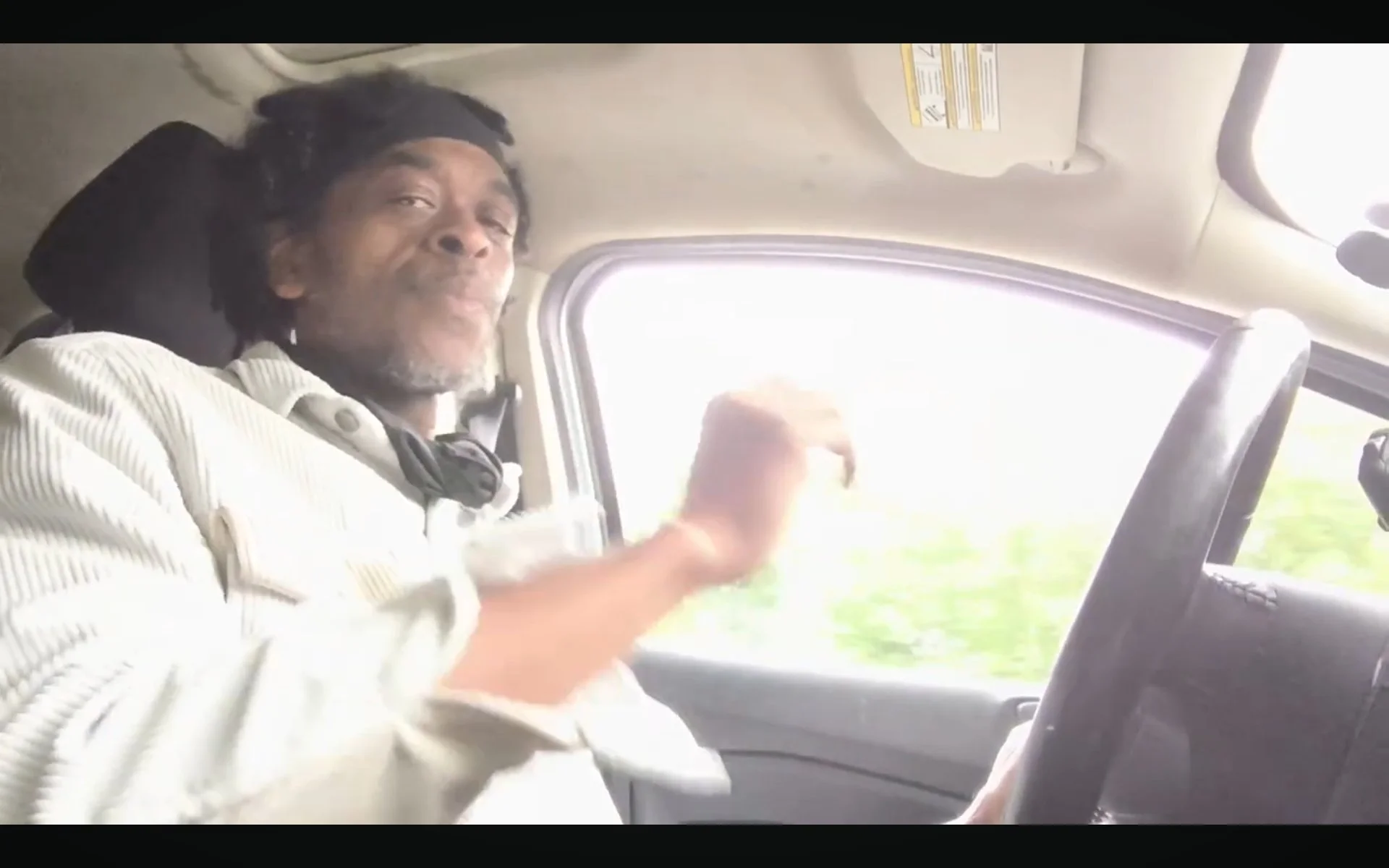 A man with dreadlocks and a beard sitting in the driver's seat of a vehicle, smiling and waving at the camera, with a bright window behind him.