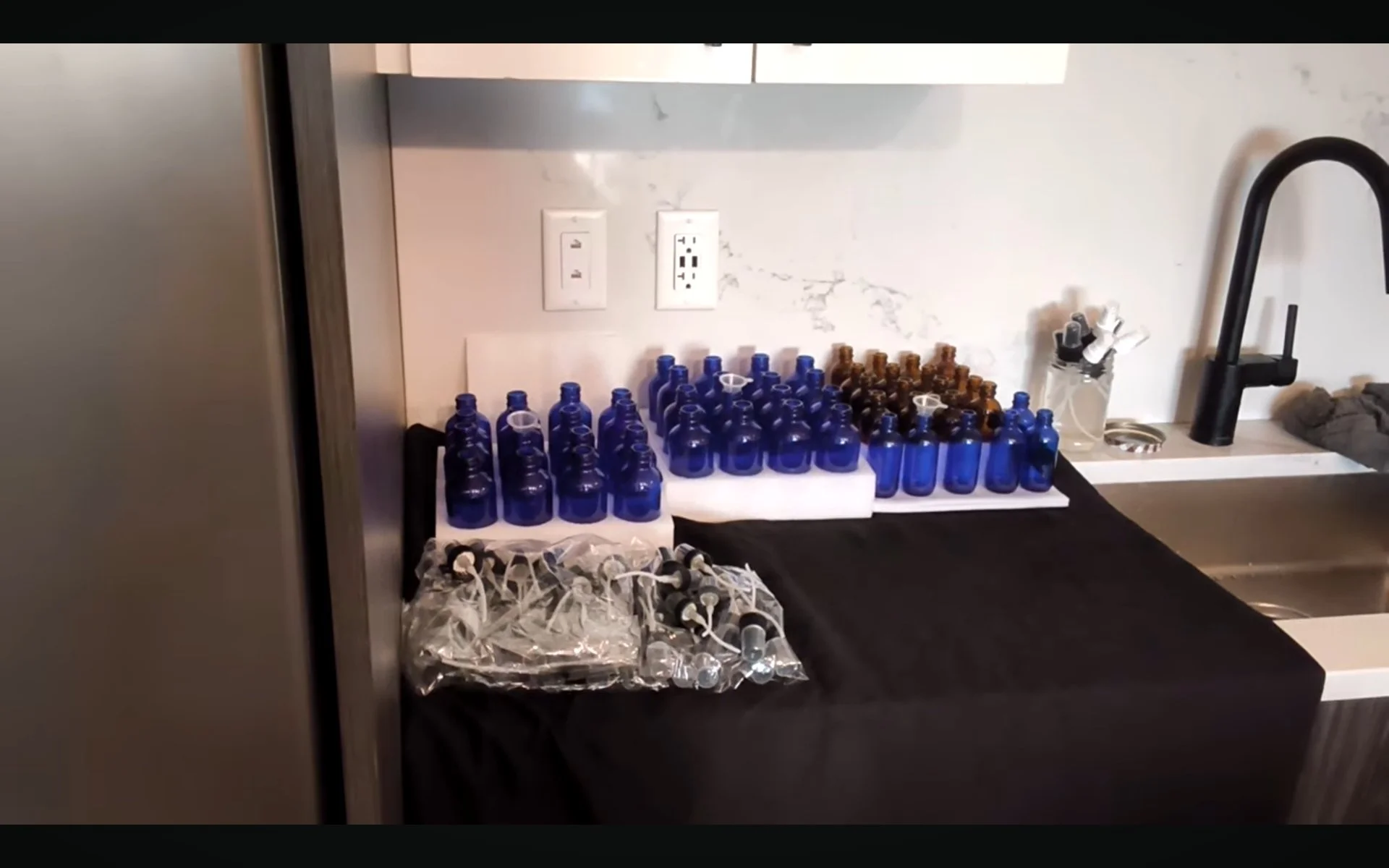A countertop with numerous small blue glass bottles, some with brown caps, arranged in clusters. A few clear plastic bags containing additional bottles are at the front. To the right, there is a sink with a black faucet and a container holding various black and white utensils, including scissors and kitchen tools. Two electrical outlets are on the wall behind the bottles, and the wall has a marble-like pattern.