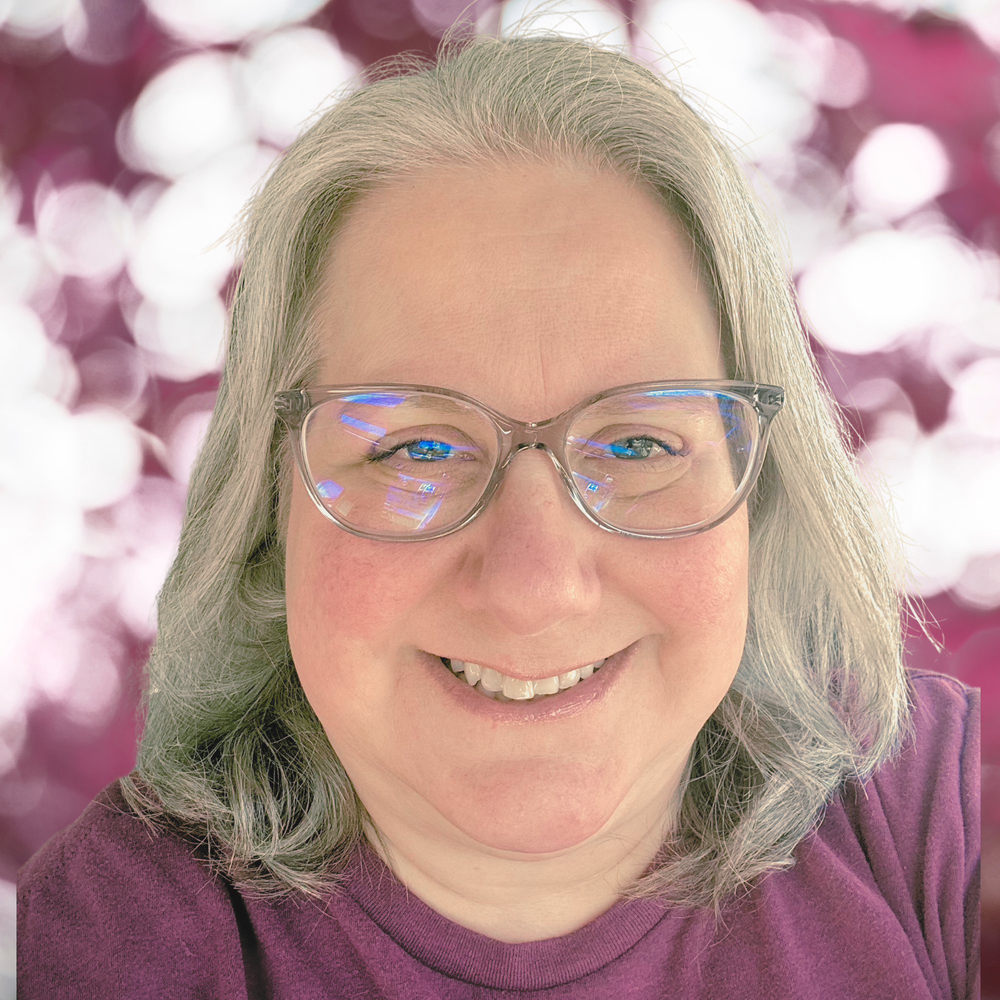 Close-up of a smiling woman with blonde hair, wearing glasses, a purple top, and a pink background with bokeh lights.