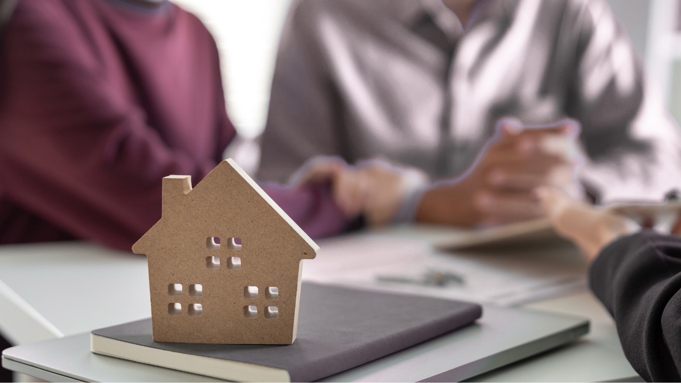 A small cardboard house model on a notebook at a table with people in the background discussing something, one person holding a phone.