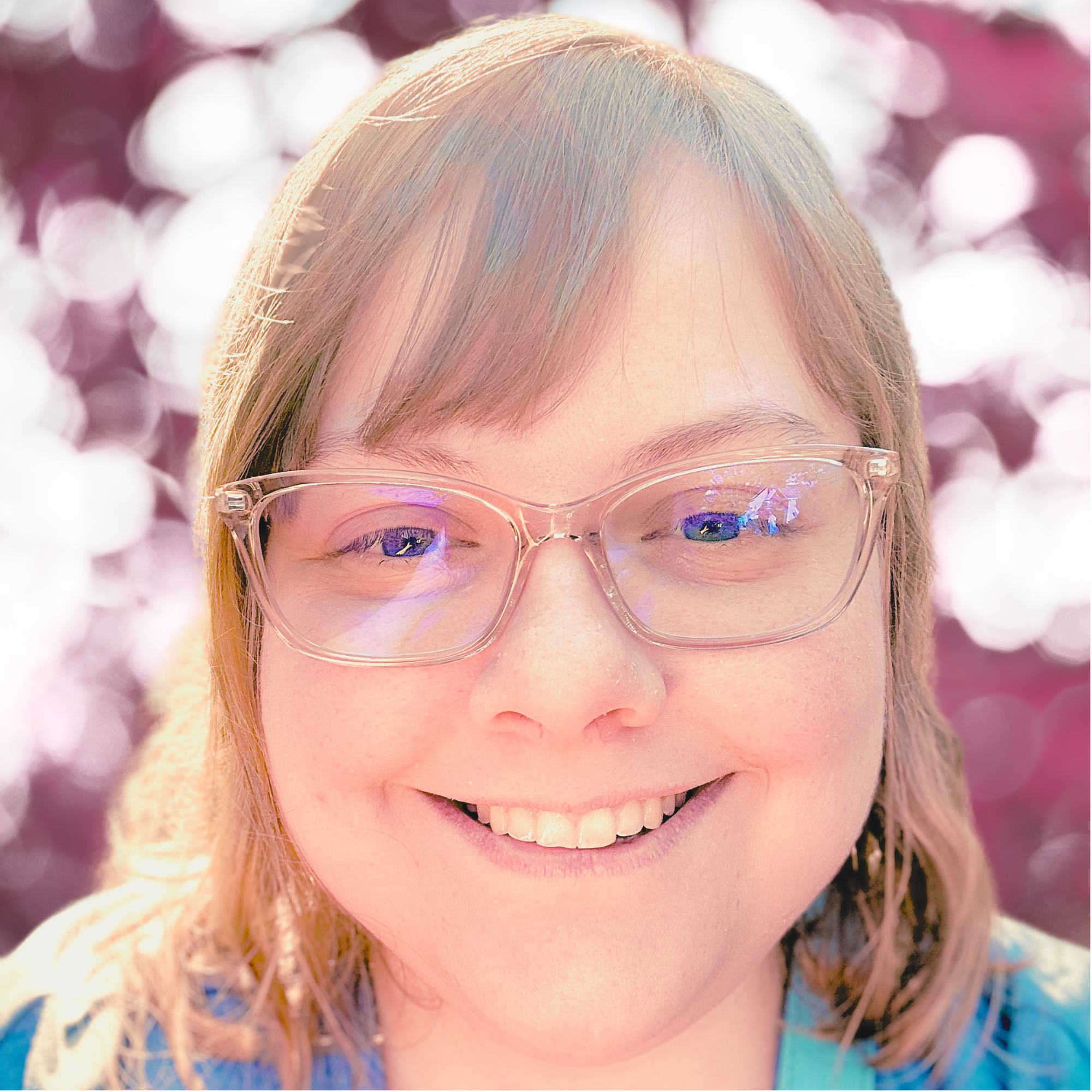 A person with blond hair, wearing clear glasses, smiling with visible teeth, with a background of pink and white bokeh lights.