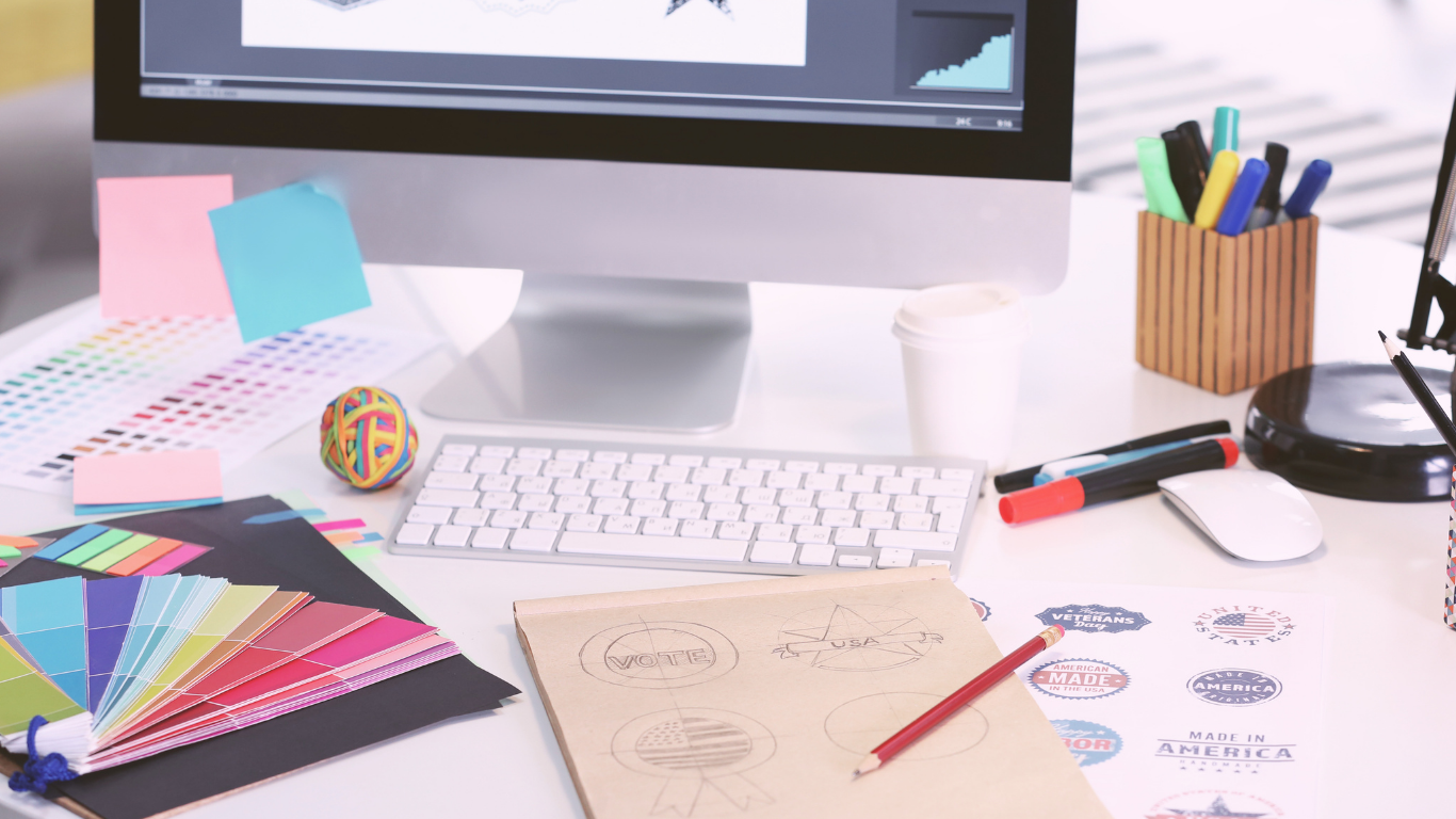 Desk with color sample book, sticky notes, a branded rubber band ball, pens, color markers, a paper cup, keyboard, mouse, a computer monitor displaying a graph, a sketchpad with designs, and stickers on the desk.