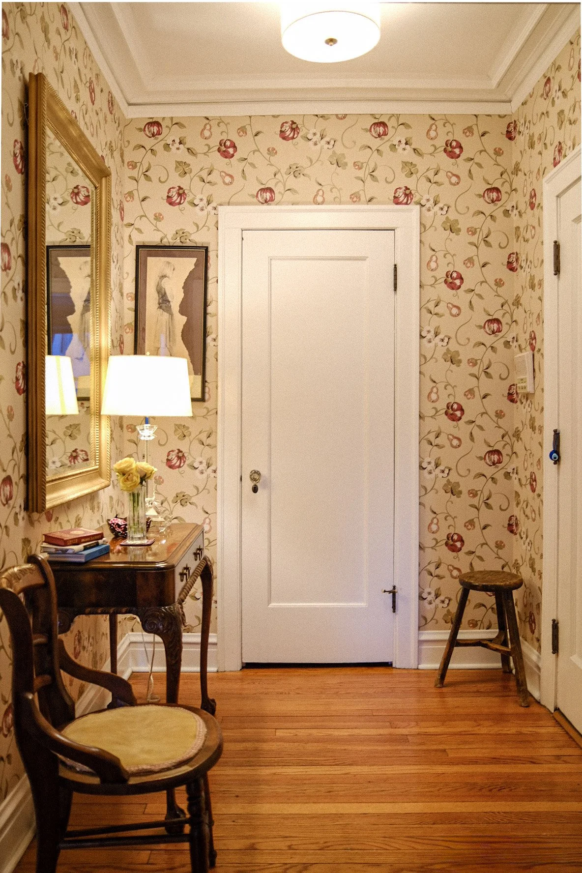 A floral patterned hallway in Lakeview East Chicago with a white door in the center, a small wooden table with a glass vase of yellow roses and a lamp, a gold-framed mirror, and a wooden stool, with hardwood flooring.