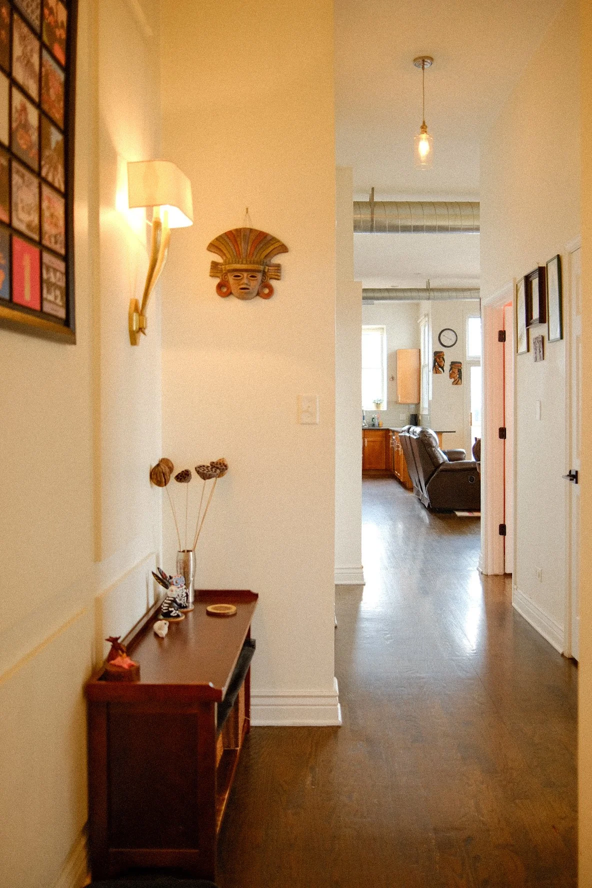 View of a hallway with wall decorations, a small table with decorative items, and a living room in the background with furniture and windows.