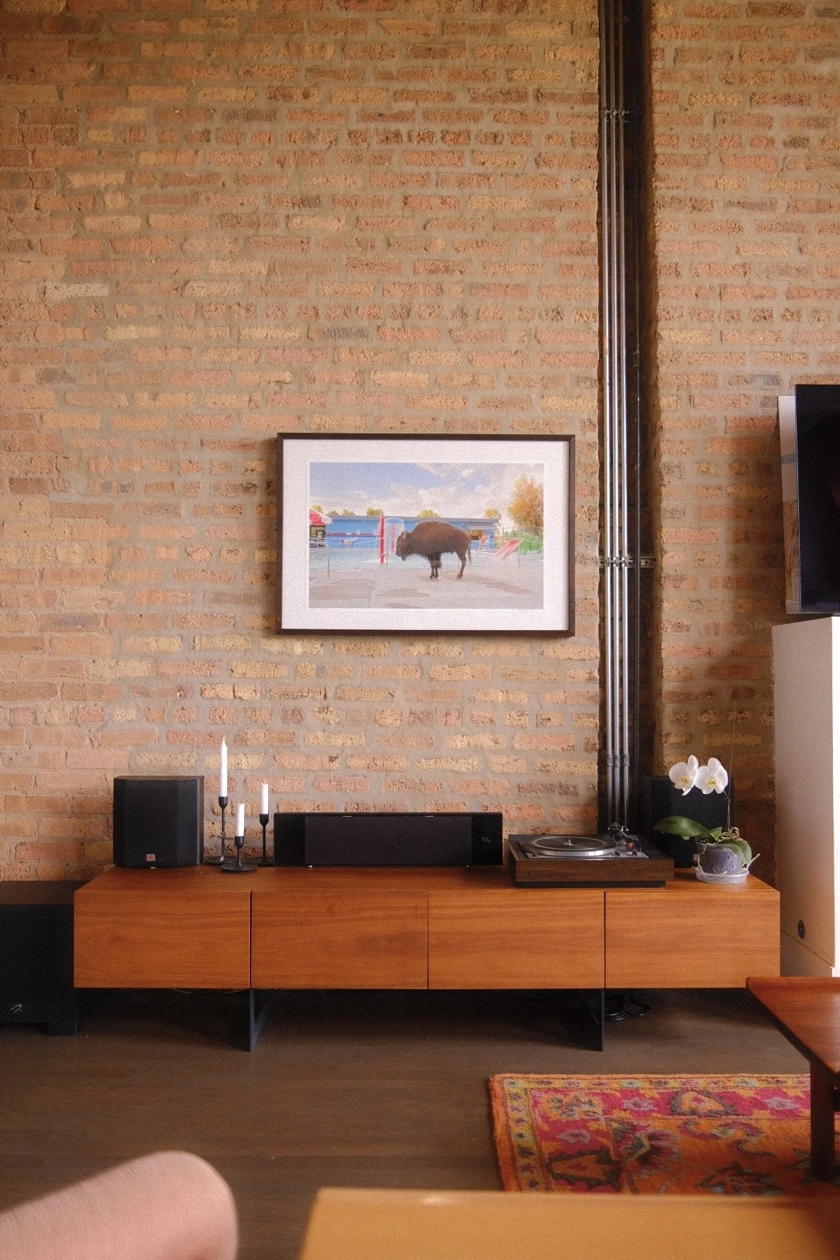 A brick wall living room in Humboldt Park, Chicago with framed photo of a bison in a playground. Below, a wooden sideboard with candles, speaker, and a turntable, accented by a potted orchid, a colorful rug on the dark wood floor.