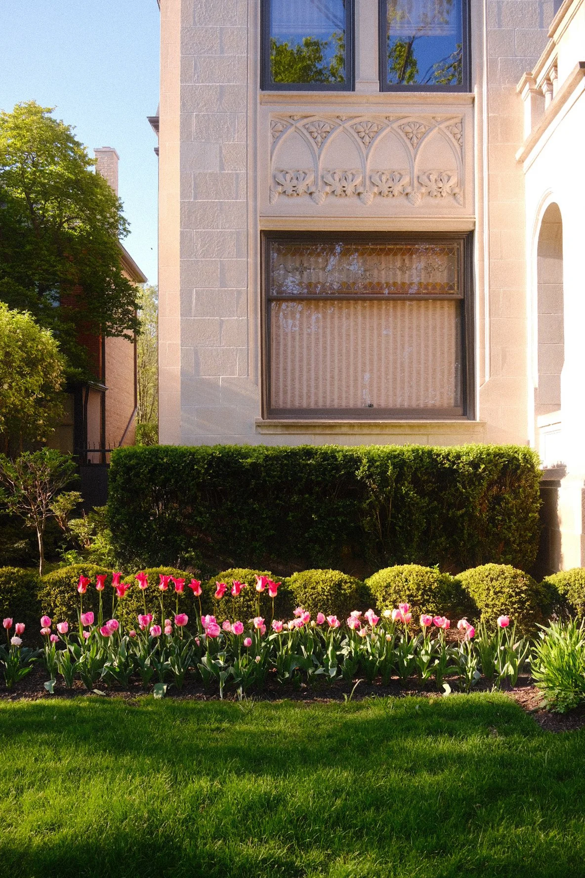Front view of a beige stone building with large windows, decorative architectural details, and a garden with pink tulips, green bushes, and a well-manicured lawn.