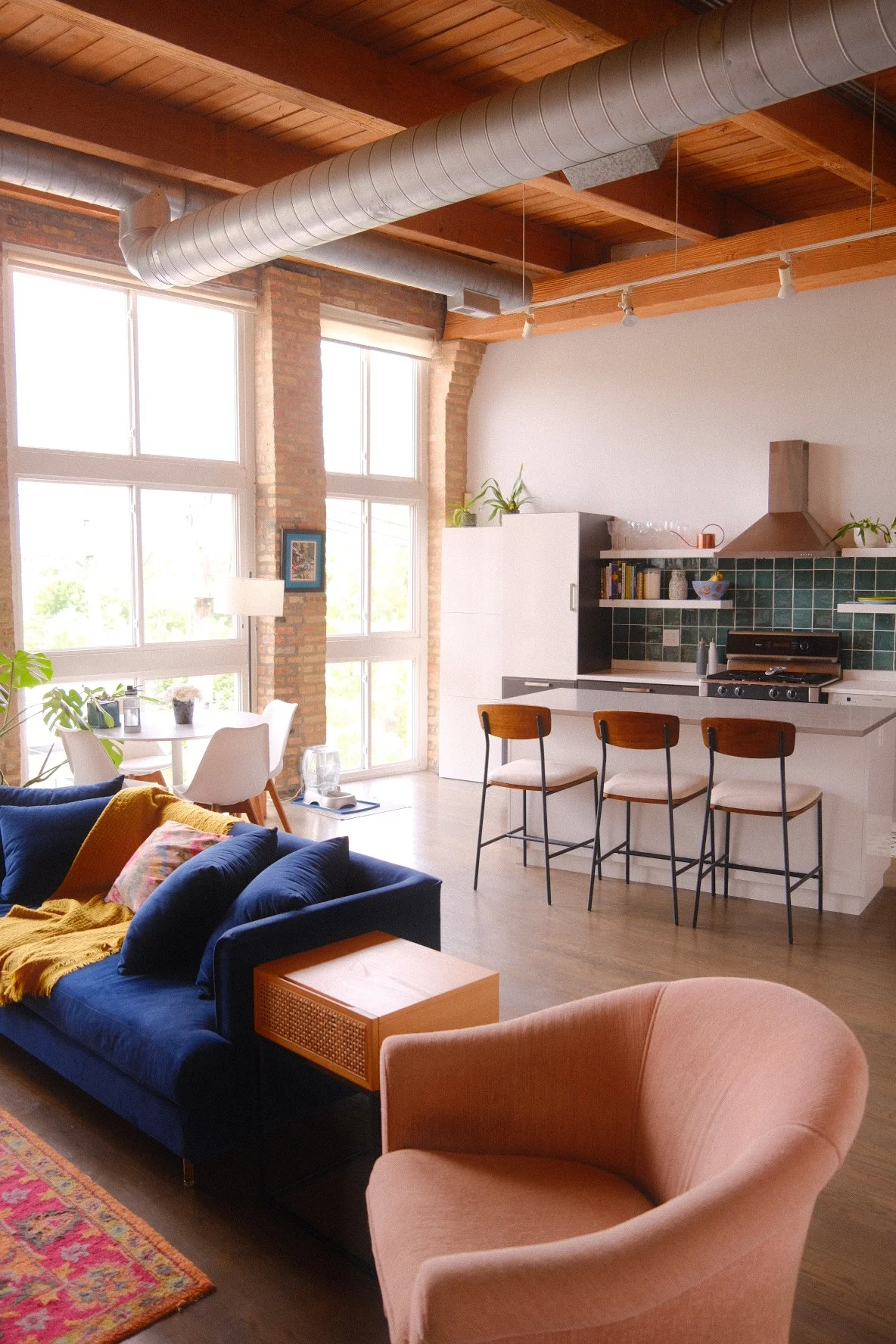 Open-concept living room in Humboldt Park Chicago and kitchen with large windows, exposed brick walls, wooden ceiling beams, modern furniture, and minimal decor.
