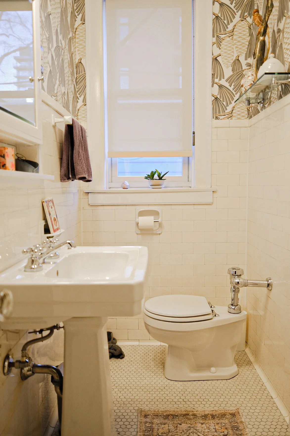 A vintage bathroom with white subway tile walls, a pedestal sink, a toilet, and a window with a roller shade. The window sill has a small potted plant and a shell. There are built-in shelves with decorative items and towels. The floor has small hexag