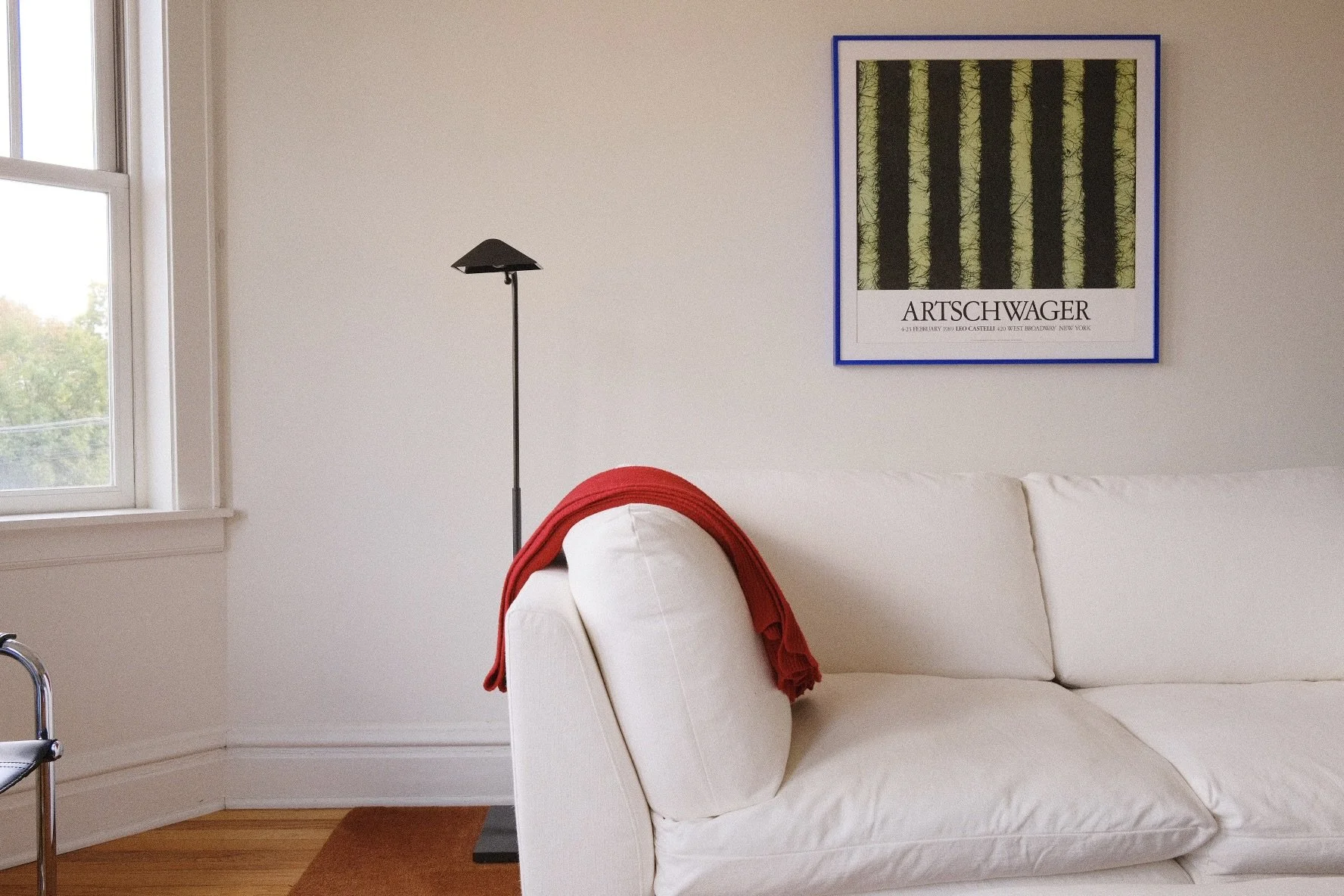 A white sofa in Wicker Park Chicago with a red blanket draped over the armrest, a black floor lamp, a window with a view of trees, and a framed art poster on the wall featuring an abstract image of green and black vertical stripes.