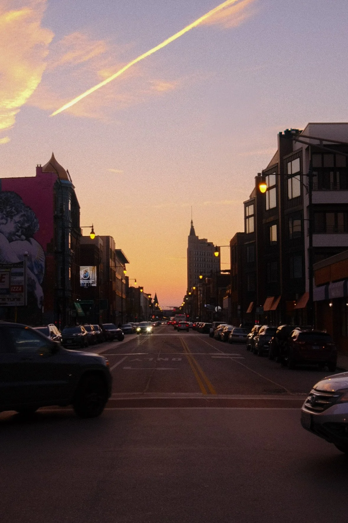 A Chicago street in Wicker Park at sunset with cars parked along the sides and buildings on both sides. The sky is pink and orange with a few clouds and a contrail.