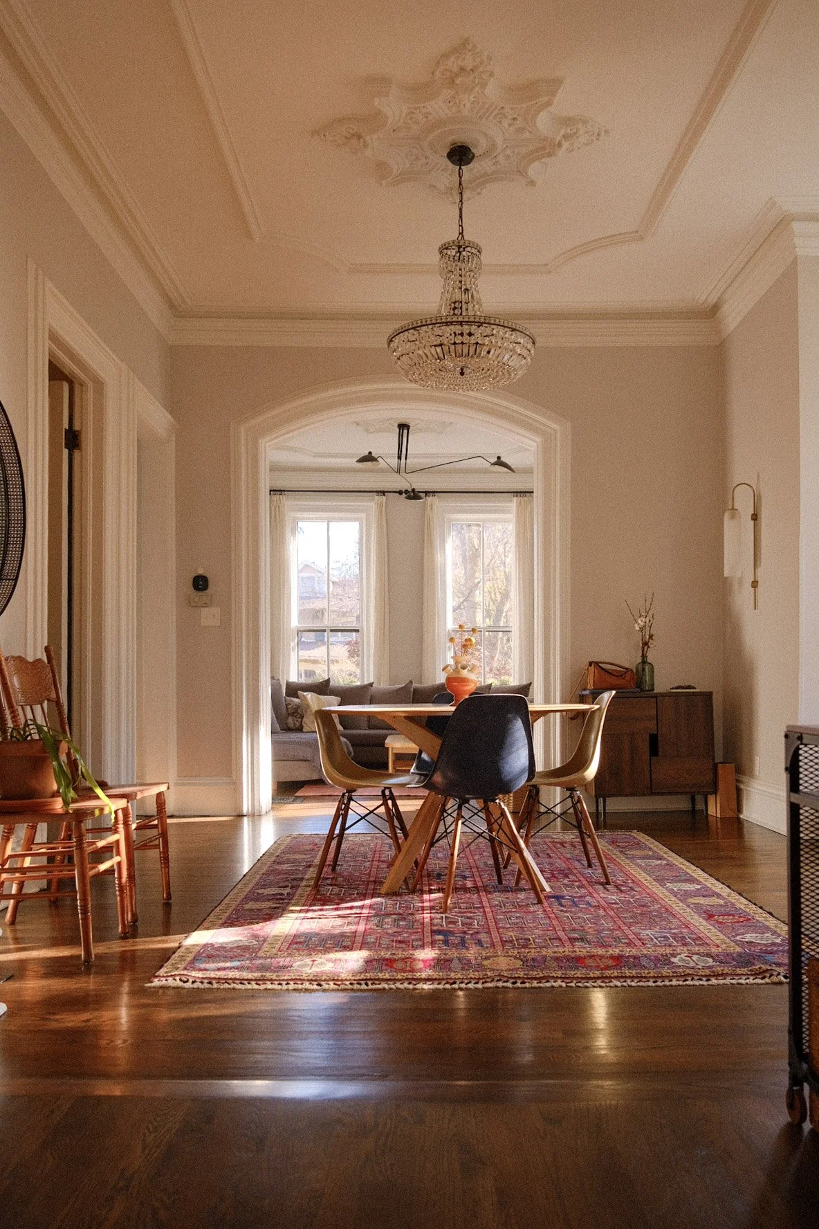 Dining room in Evanston with wooden table and chairs on a colorful rug, a chandelier hanging from ornate ceiling, and a view of a cozy living area with a sofa, large windows, and decorative items.