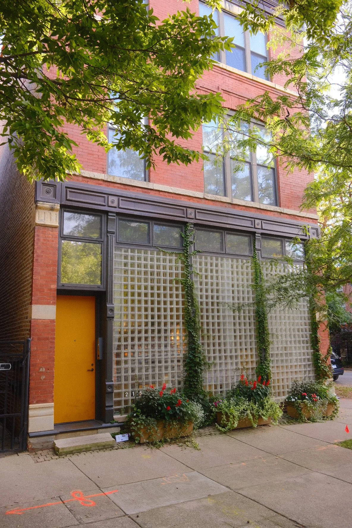 The image depicts a multi-story building with large windows on a brick facade. The ground floor has a prominent yellow door and a decorative glass block wall with climbing plants. There are flower pots with colorful flowers on the sidewalk in front o