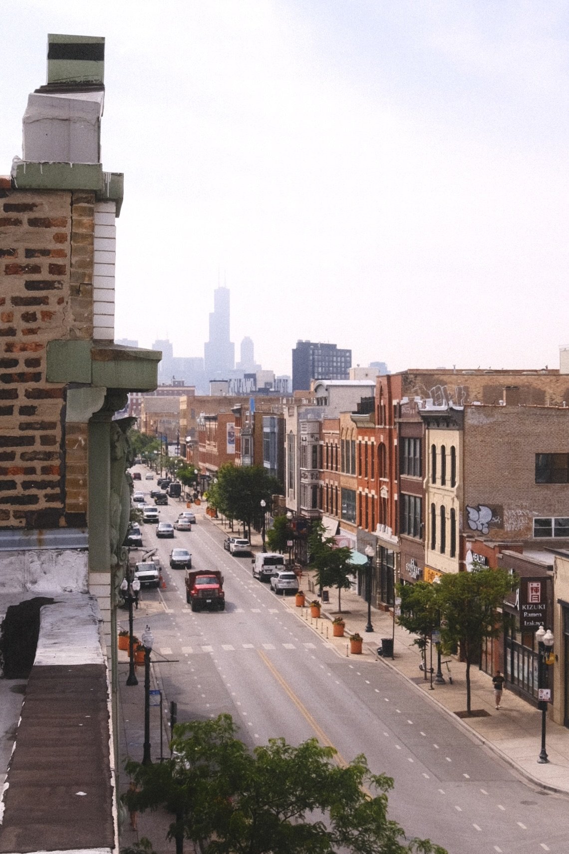 A city street view with buildings on both sides, parked cars, and a skyline with the Willis Tower in the distance. Wicker Park Chicago.