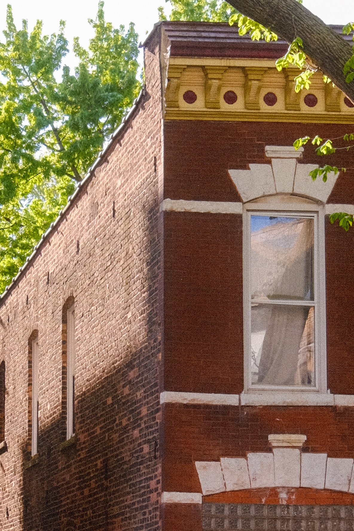 Close-up of the upper corner of a brick building with decorative architectural details and a window, with a large tree with green leaves in the background.