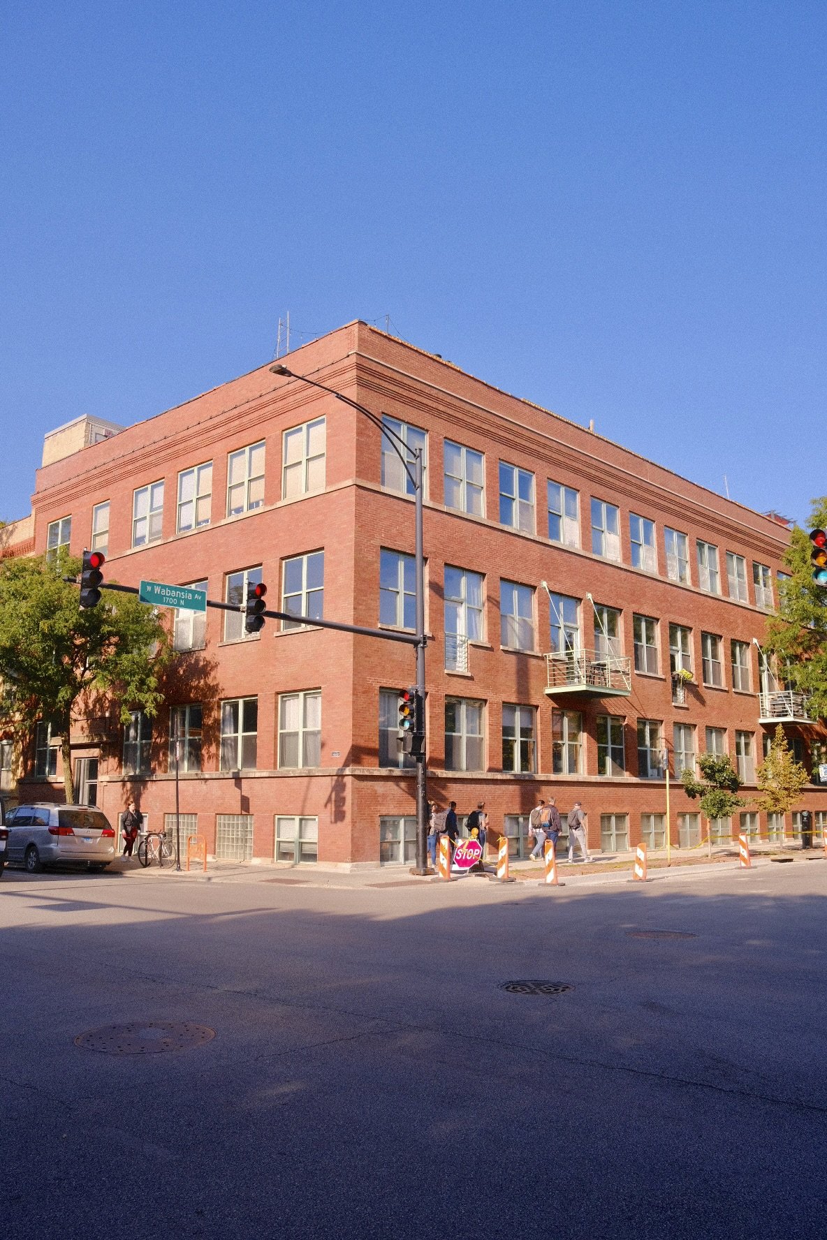 A red brick loft condo building in Bucktown Chicago on a street corner under a clear blue sky, with pedestrians and cars nearby. Street signs indicate W Wabansia Ave, 1700 N.