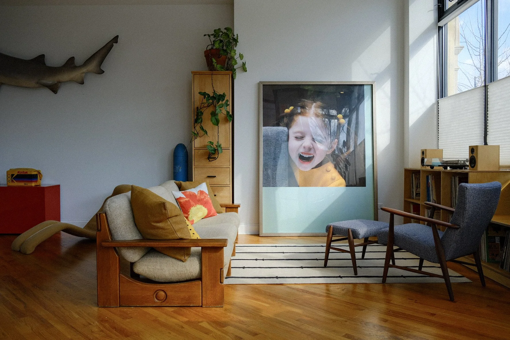 Living room in Pilsen neighborhood, Chicago with beige couch, blue armchair, and wooden furniture. Large framed photo of a girl in an orange shirt with pigtails. Sunlight streams in through tall windows.