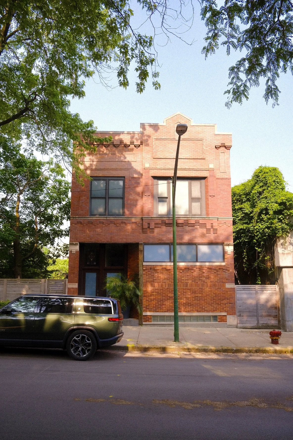 Three-story brick building on a city street in Bucktown Chicago, with a black SUV parked in front, and green trees surrounding it, under a clear blue sky.
