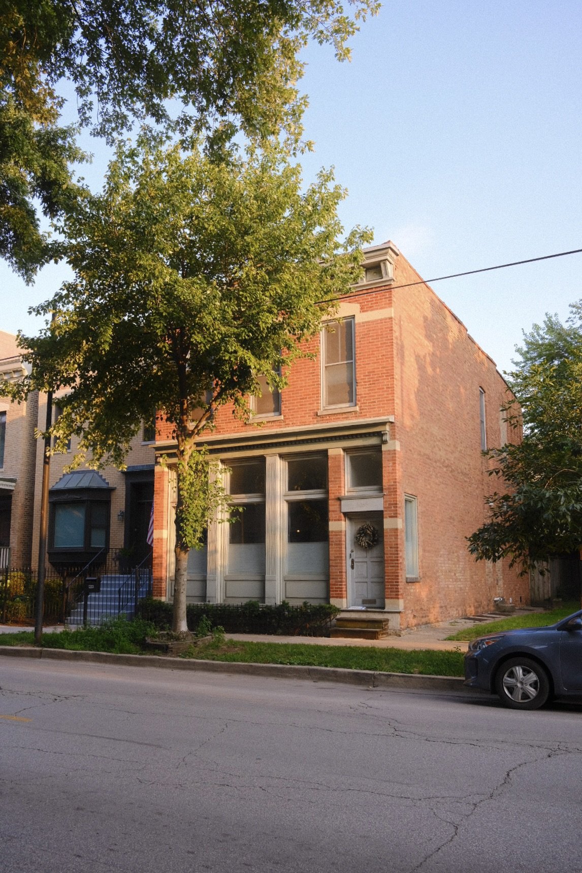 A brick townhouse in Lincoln Park Chicago with a porch and three large windows facing the street, flanked by trees, with a car parked in front on the street.