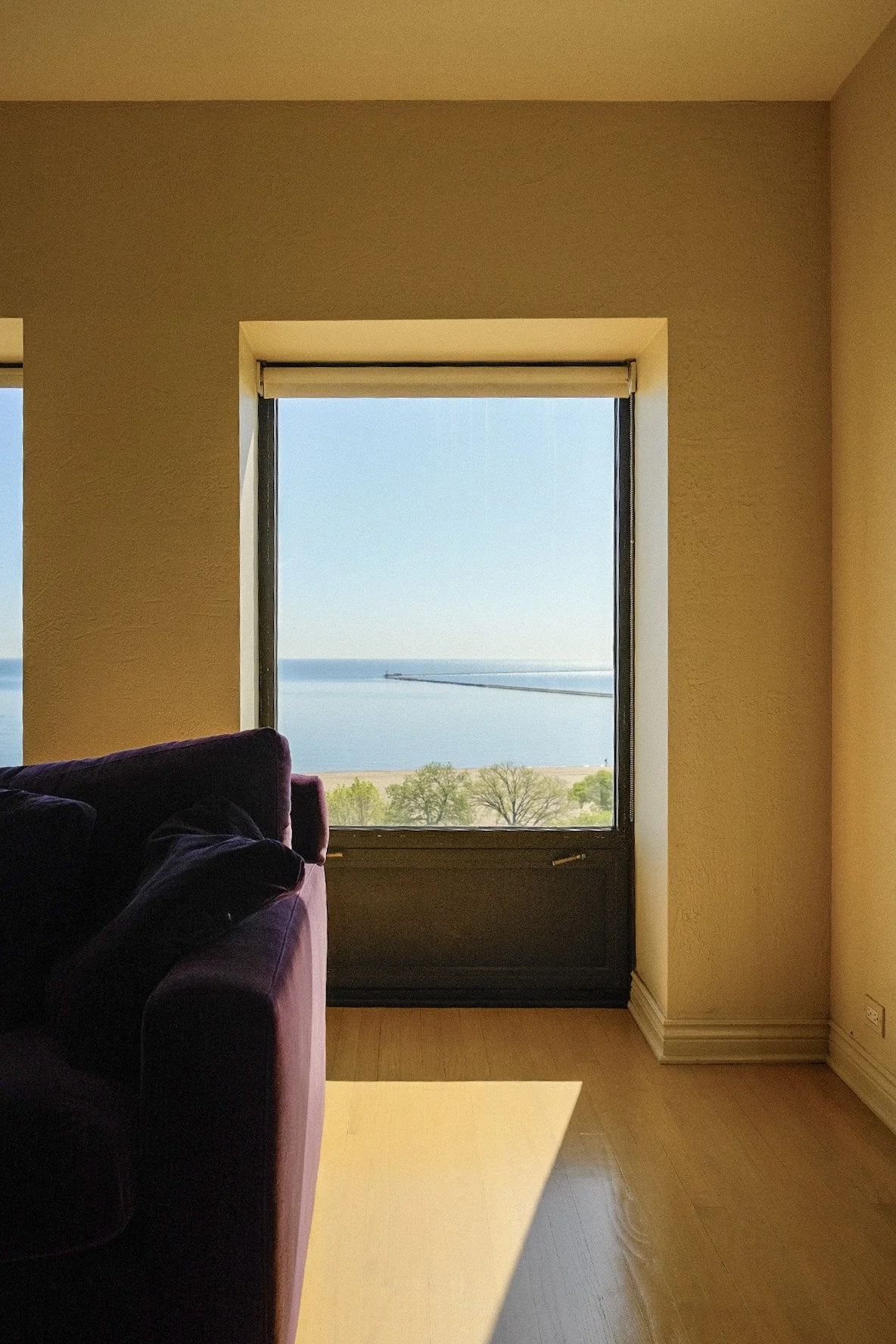 Condo living room in Gold Coast with purple sofa and large window showing a view of water, trees, and sky.