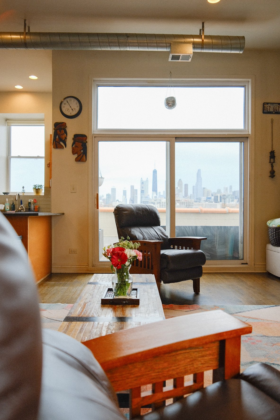 Living room in West Town Chicago with a view of the Chicago skyline through large windows, a black leather chair, a wooden coffee table with a vase of flowers, and decorative wall art.