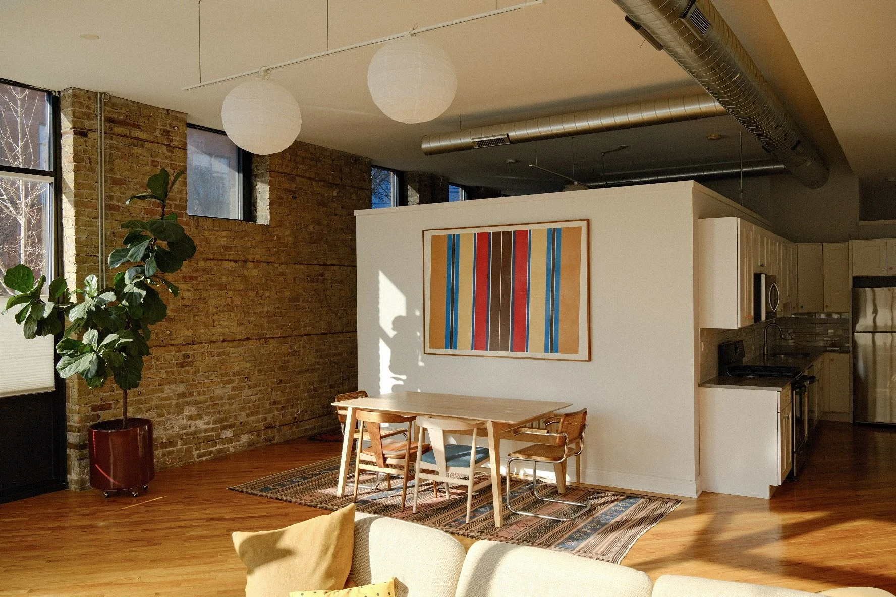 Modern open-plan living space in Pilsen, Chicago with exposed brick wall, large windows, a potted plant, a dining table with chairs, and a kitchen area with white cabinets, stainless steel appliance, and colorful wall art.