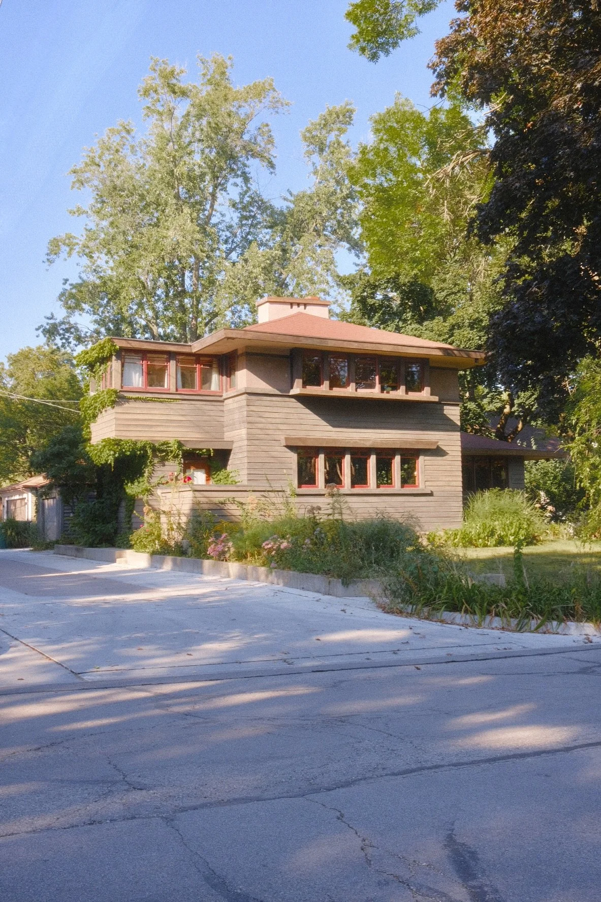 A two-story house in Oak Park with dark brown wood siding, multiple windows with red trim, surrounded by greenery and trees, with a concrete driveway in the front.