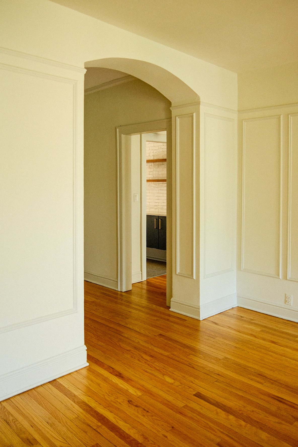 Interior view of a room in Evanston with hardwood floors, white paneled walls, and an arched doorway leading to a kitchen area with white brick walls, wooden shelves, and black cabinets.