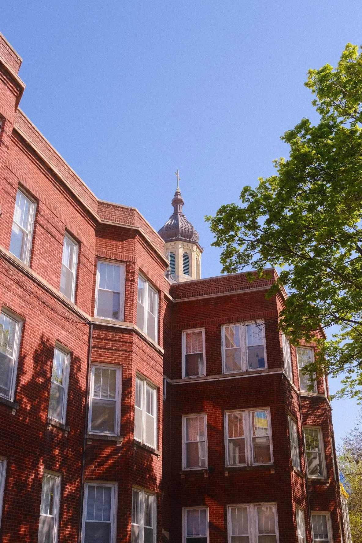 A red brick apartment building in Ukrainian Village Chicago with multiple windows and a tree on the right side, with a church steeple topped with a cross in the background against a clear blue sky.