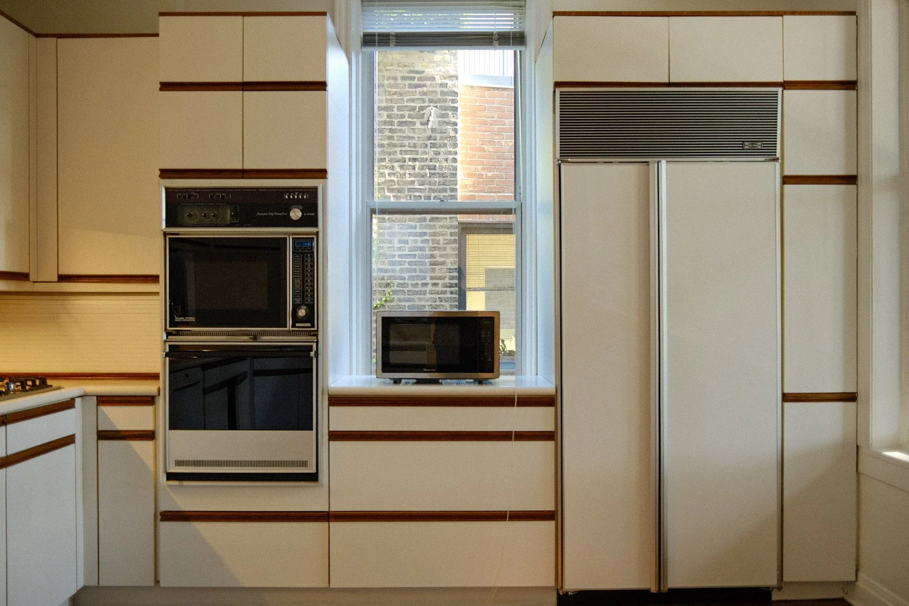 Kitchen with white cabinets and brown trim, a large window overlooking a brick wall outside, a microwave on the counter, and a small microwave on the windowsill.