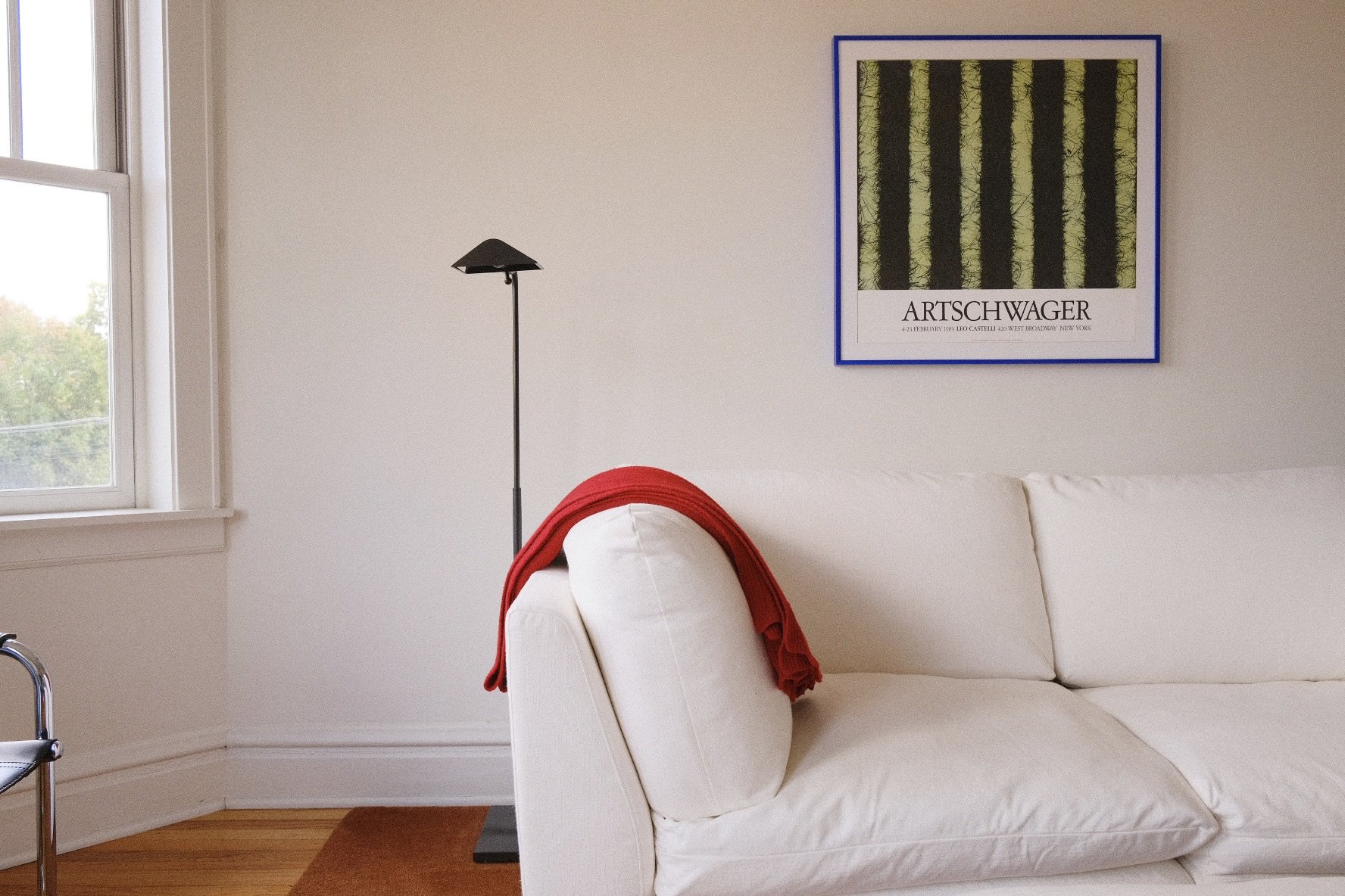 A living room corner with a white couch, a black floor lamp, a framed art poster on the wall, a red blanket draped over the couch, and a window showing trees outside.