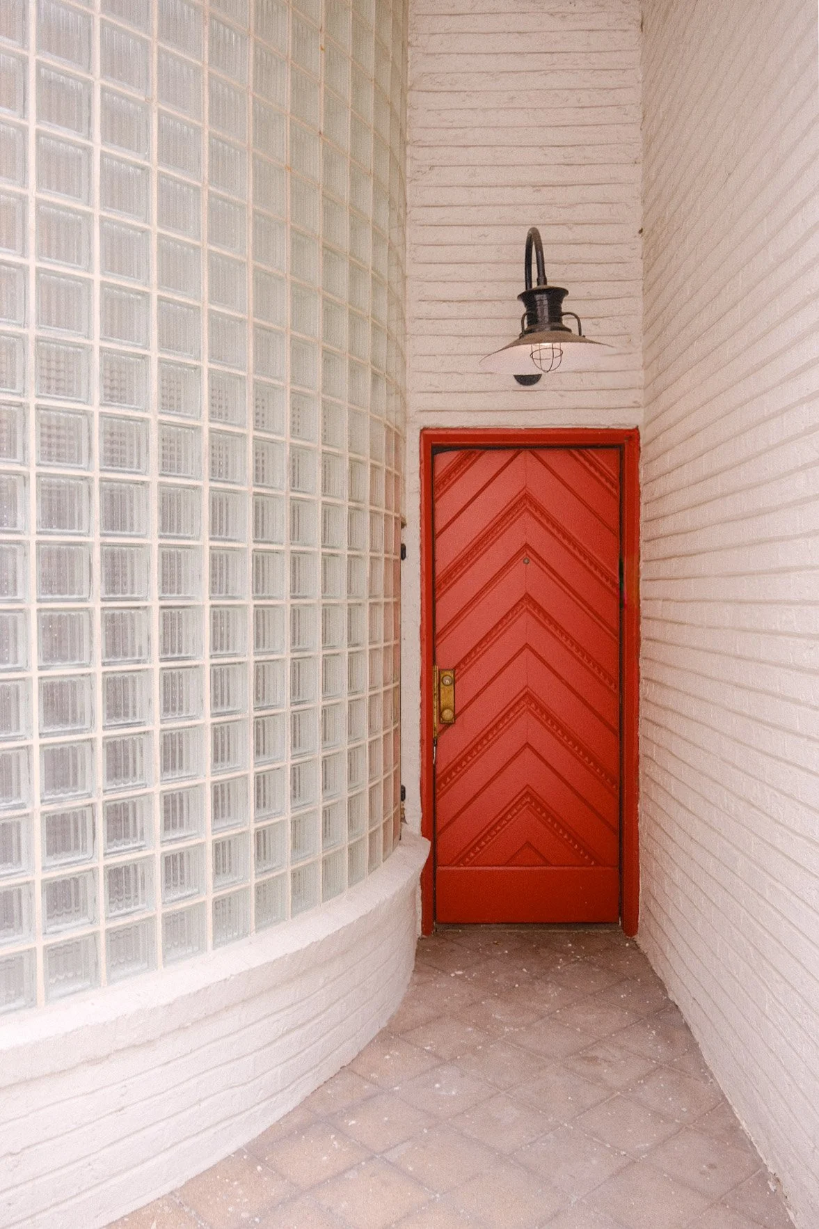 Red door with a diagonal pattern in Gold Coast, surrounded by white brick walls on either side, with a glass block wall on the left side and an industrial-style wall sconce above. Edgar Miller design.