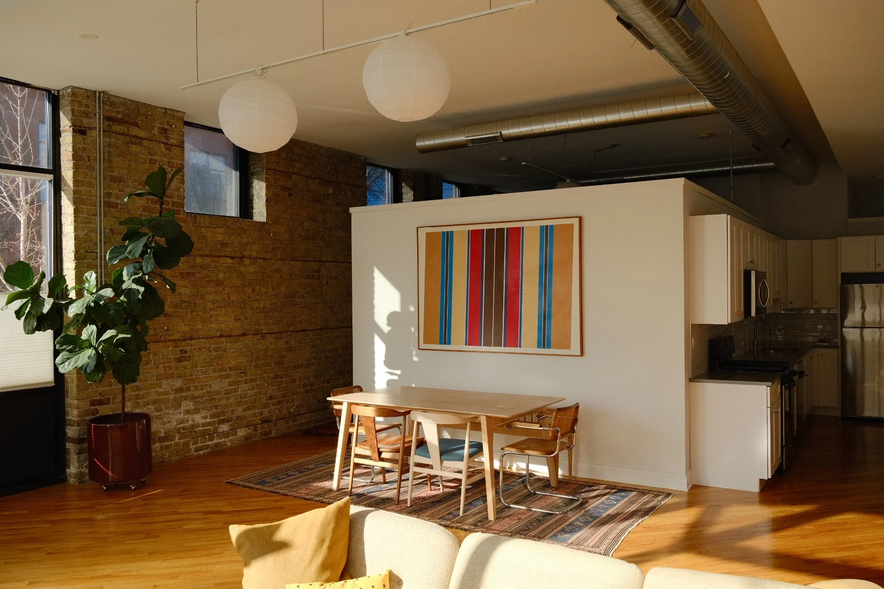 Modern apartment interior with an exposed brick wall, large windows, and a white partition with colorful striped artwork. There is a wooden dining table with chairs, a potted plant in a red pot, and white hanging paper lanterns on the ceiling.