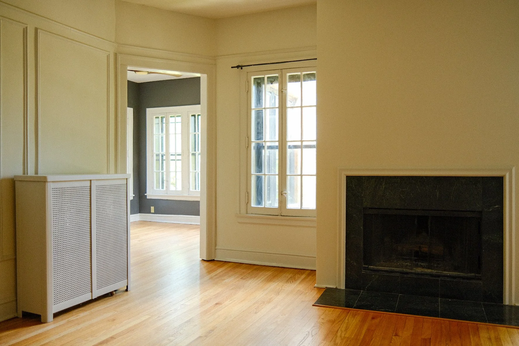 Interior of a house with hardwood floors, cream-colored walls, a black marble fireplace, and large windows with multiple panes, allowing natural light to fill the room.