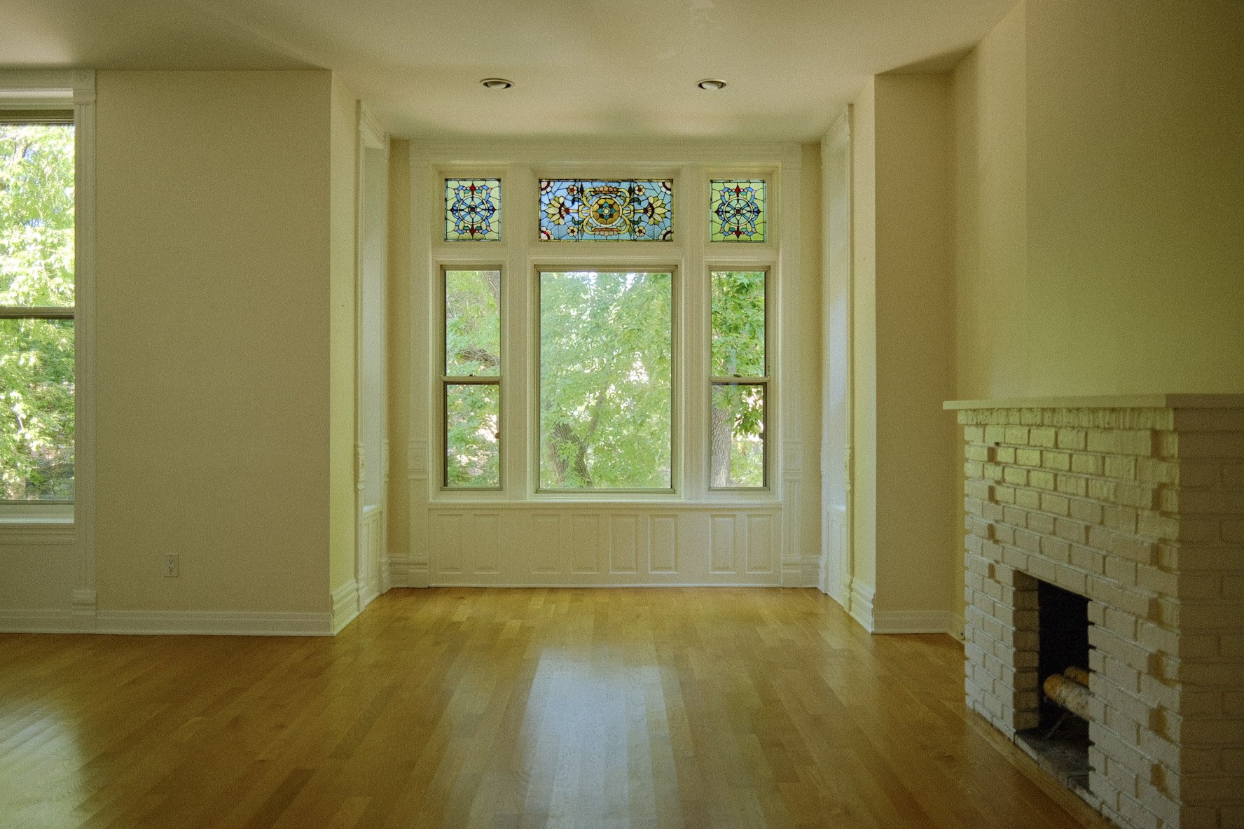 A bright, empty living room with large windows, a white brick fireplace, hardwood floors, and cream-colored walls.