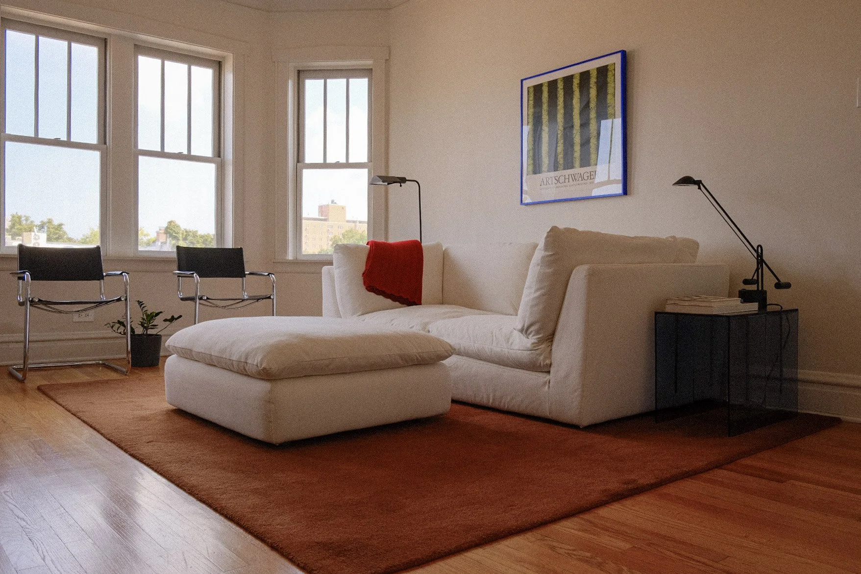 A living room with a white sectional sofa, a red throw blanket, and a black side table with books and a modern black desk lamp. Two black chairs are near tall windows with views of buildings and trees. A framed artwork with green and yellow patterns 