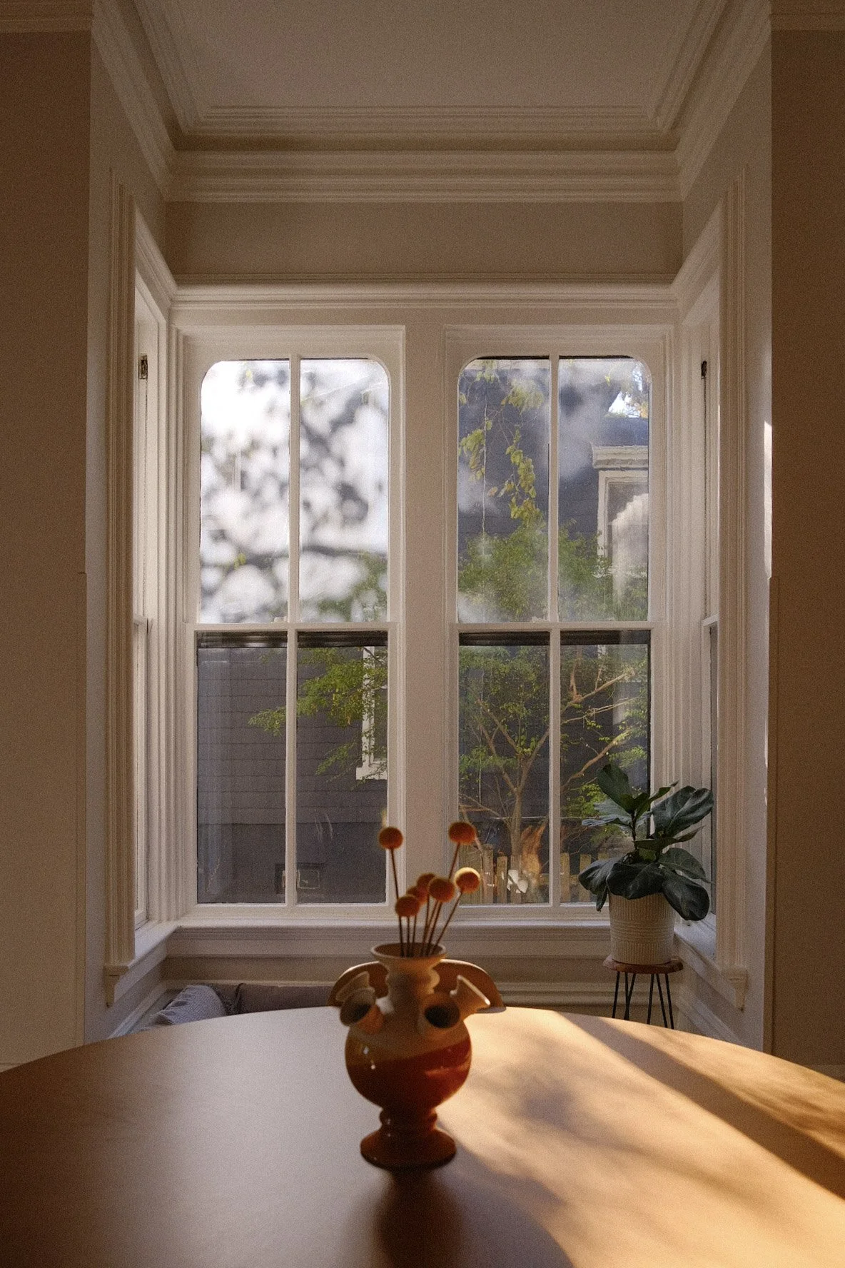 A cozy indoor space with a large, decorative window revealing trees outside. Inside, there is a table with a decorative vase holding flowers, and a potted plant on a small stand near the window.