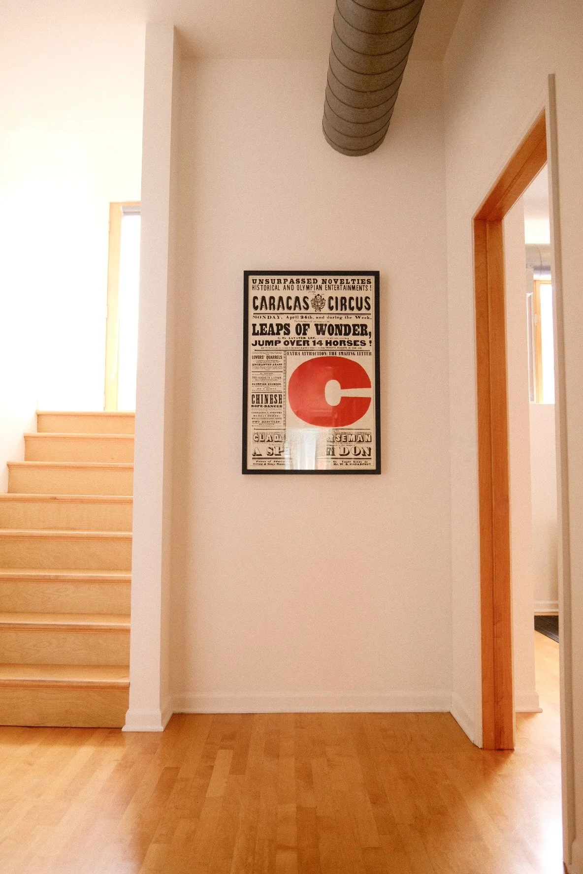 Interior view of a hallway showing a framed poster on a white wall. The poster advertises a circus event with bold text and graphic elements, including a large red circle with a white letter 'C' in the center. To the left, there is a staircase with l
