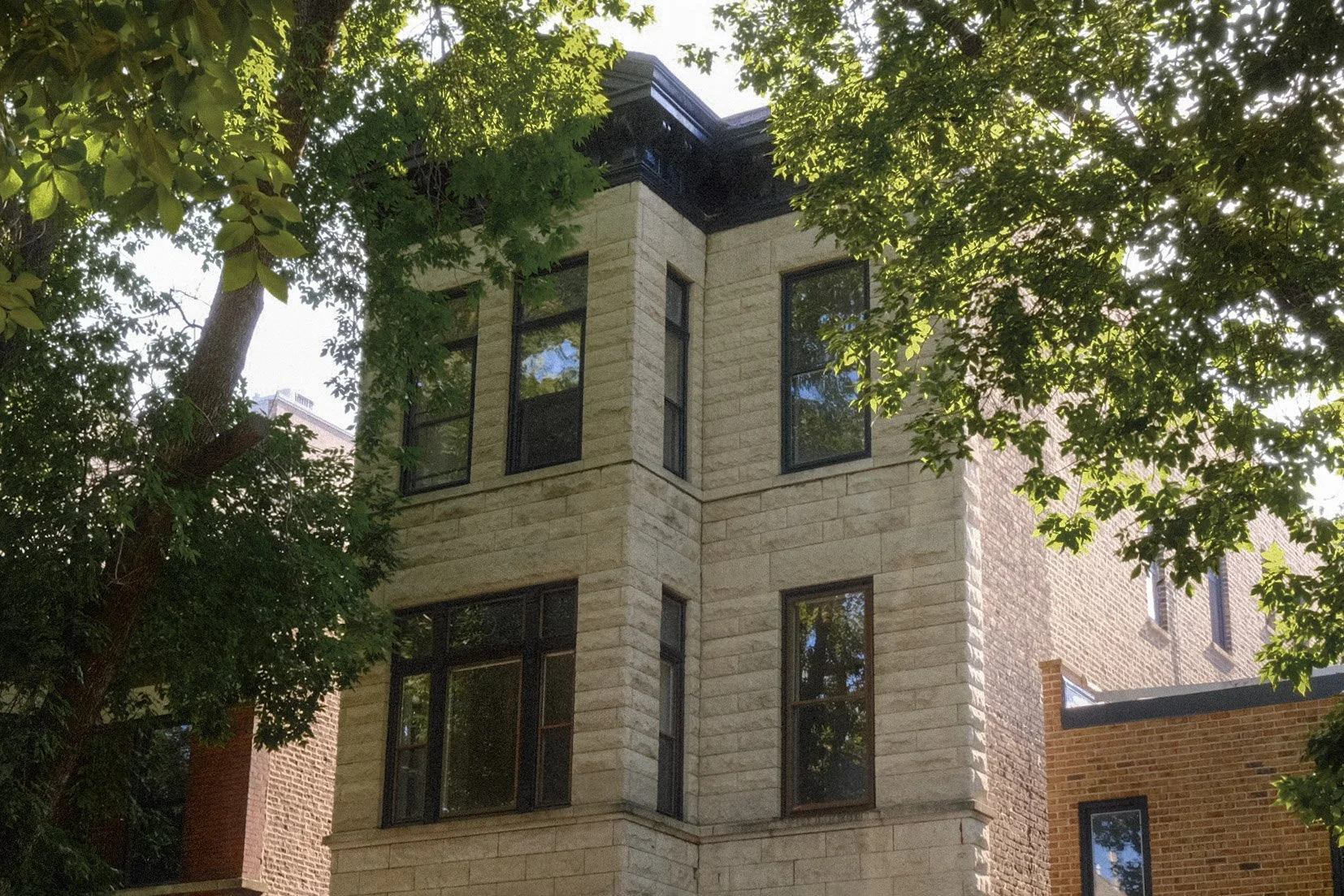 A multi-story building with beige stone exterior walls and large rectangular black-framed windows, partially obscured by leafy green trees in the foreground.