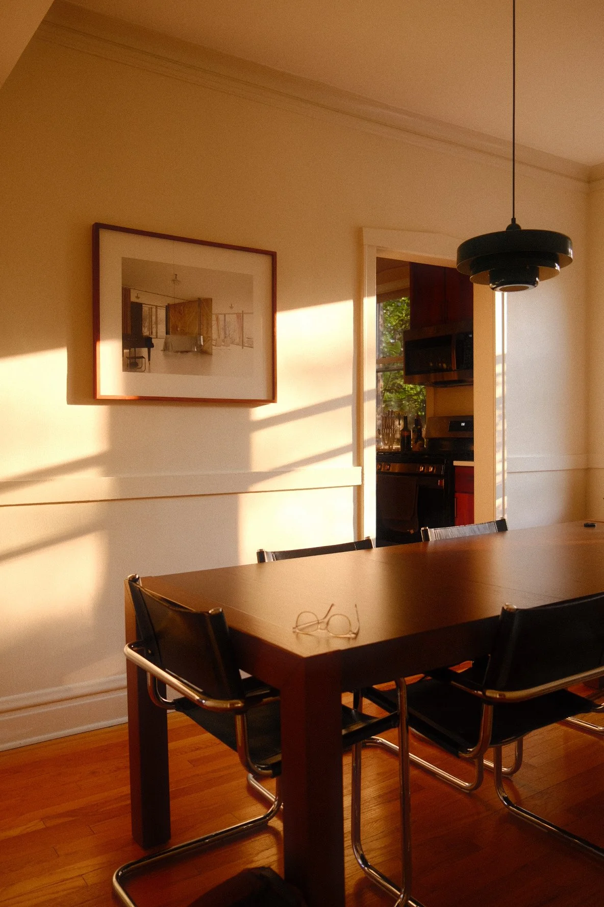A dining room in Wicker Park, Chicago with a wooden table, black chairs, a pair of glasses on the table, a framed picture on the wall, and sunlight streaming through the window into the kitchen.