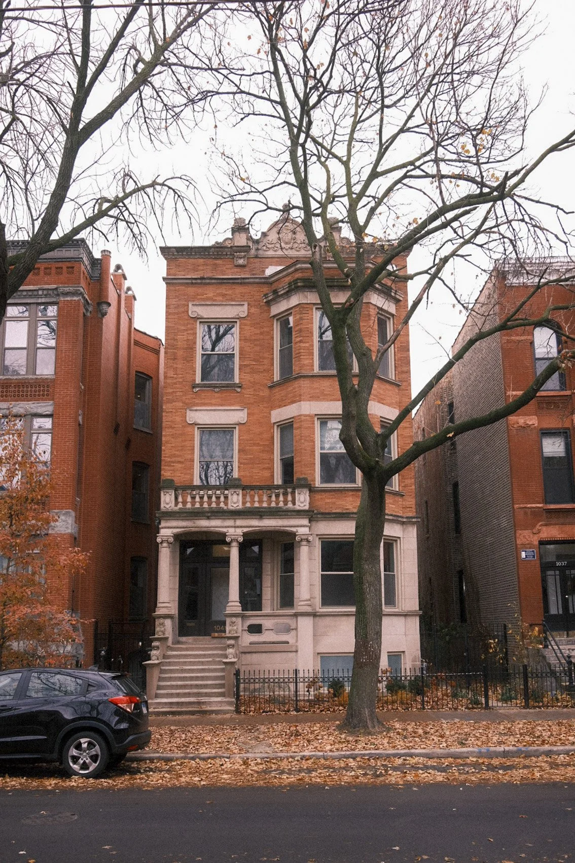 A multi-story red brick residential building with decorative stone details and a small front porch, centered amidst leafless trees on a cloudy day, with fallen leaves on the sidewalk and parked cars in front.