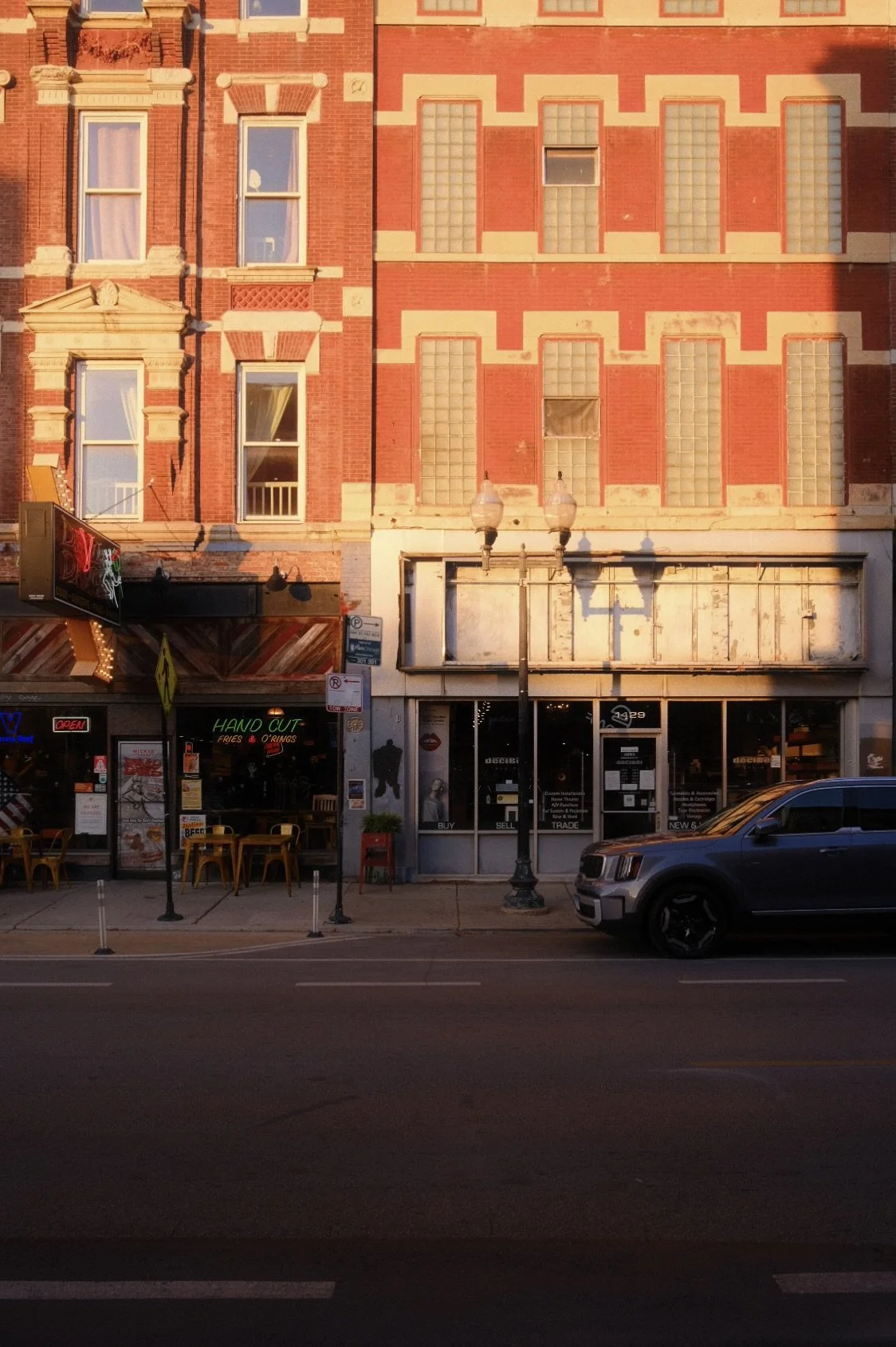 A city street scene featuring a brick building with multiple windows, some with curtains, and ground-floor storefronts including a restaurant with outdoor seating and a neon sign that reads 'Hand Cut Fries & Rings'. There are parked cars, street lamp